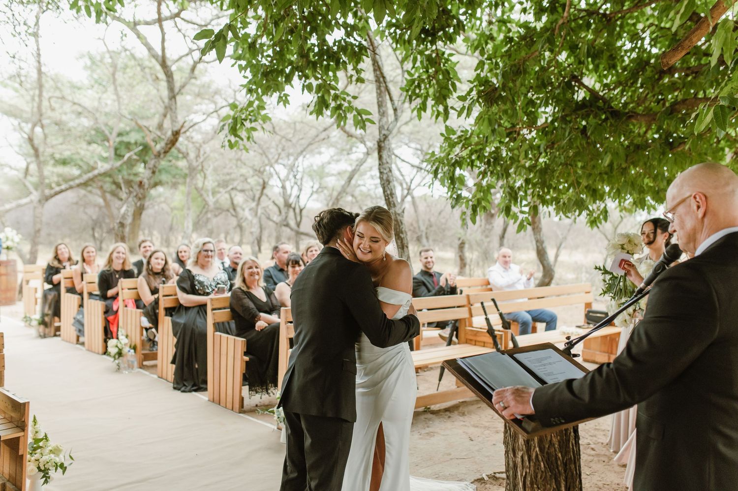 Wedding ceremony takes place in natural setting with wooden seating and overhanging tree branches.