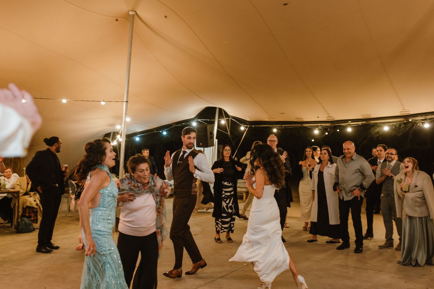 Guests dance and celebrate at a tented wedding reception with string lights creating a warm atmosphere.
