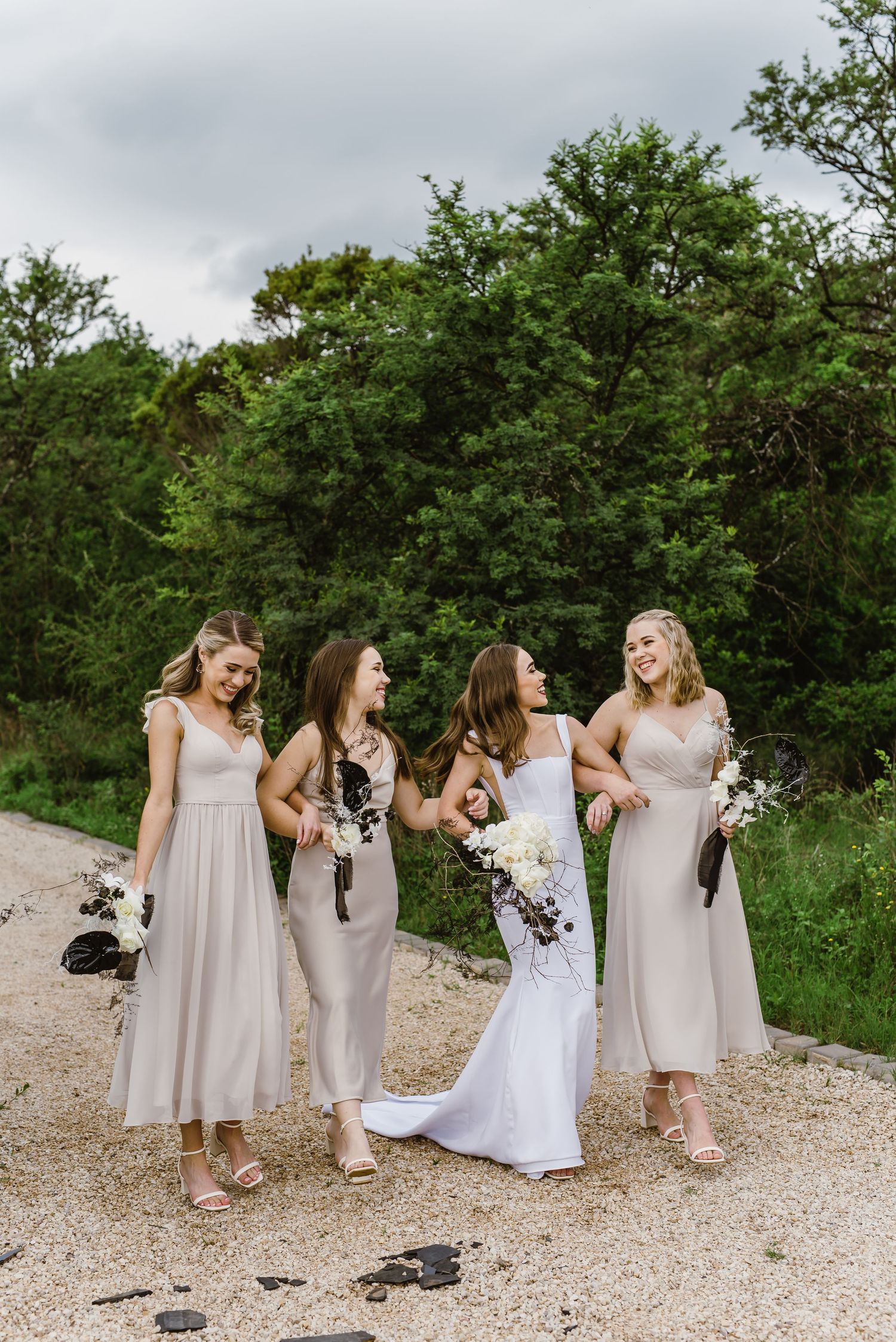 Wedding party in neutral dresses poses casually on gravel path surrounded by green foliage.