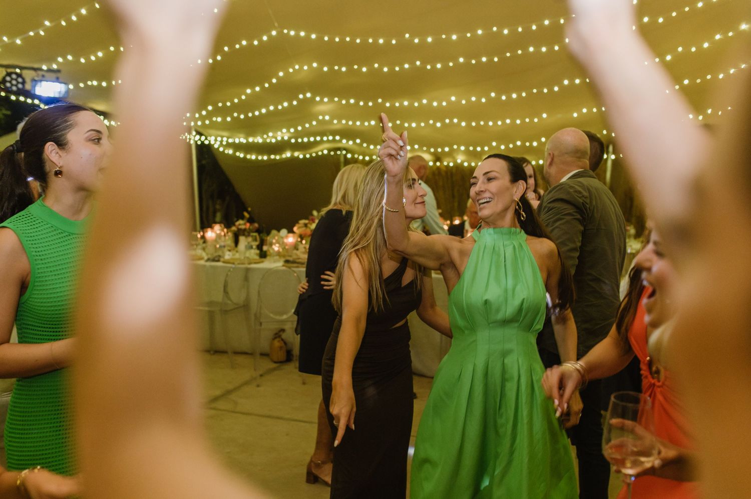 Wedding guests dancing and mingling beneath glowing string lights at an indoor reception venue.