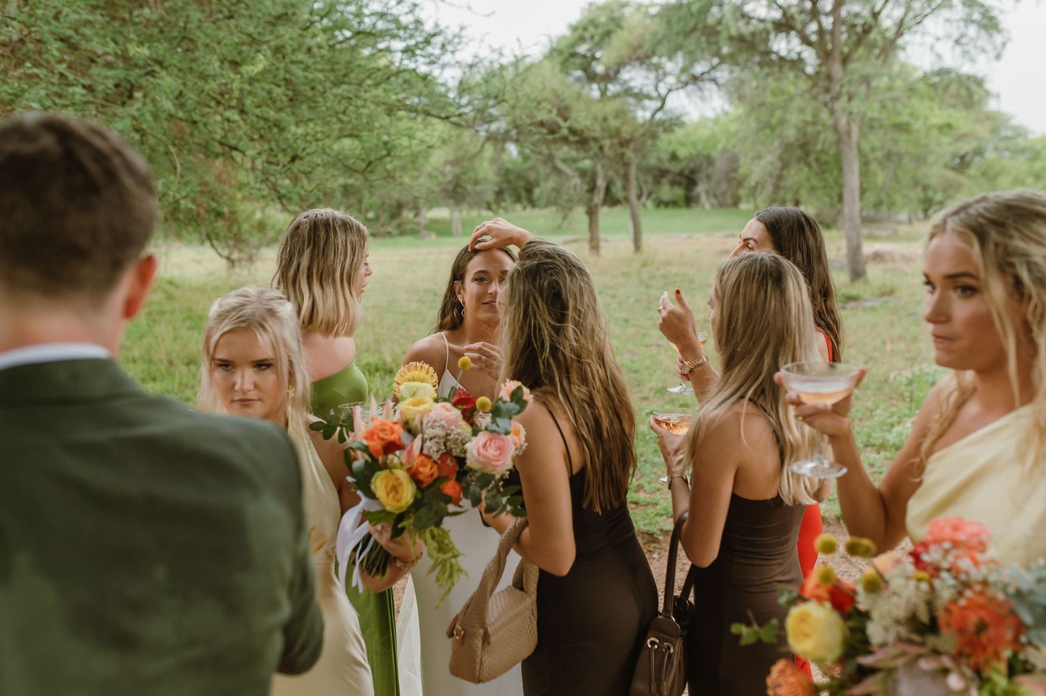 Wedding party gathered outdoors under trees holding colorful bouquets in warm natural lighting.