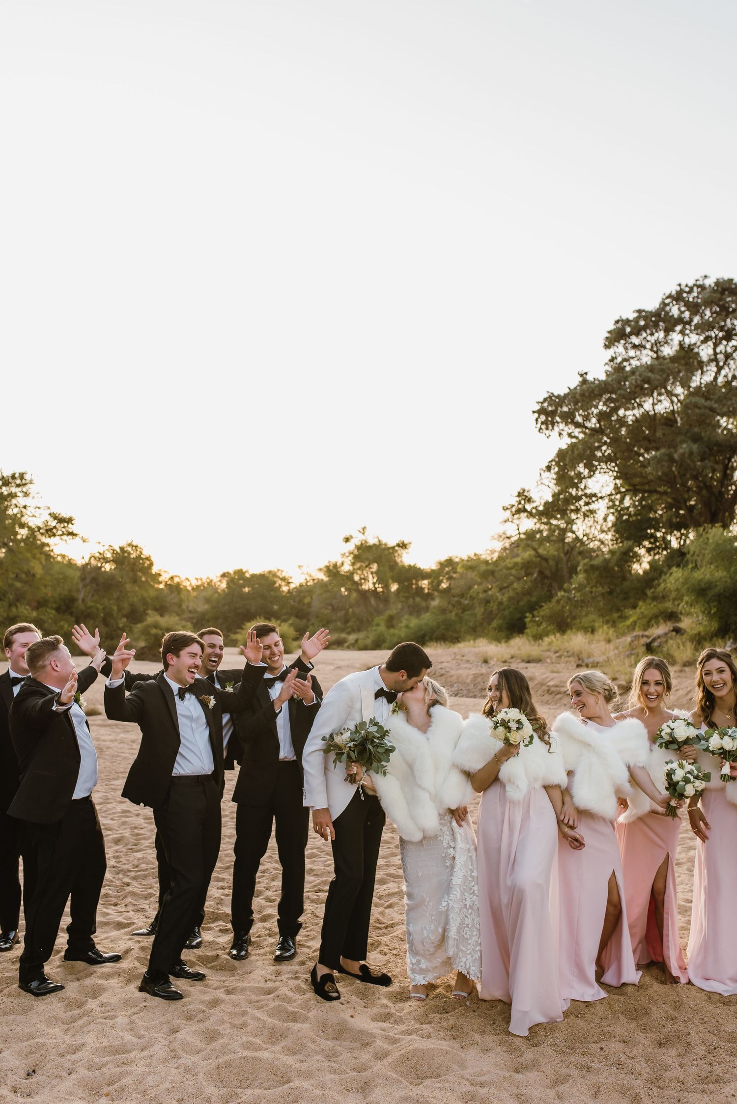 Wedding party members interact playfully while lined up for group photos on beach.