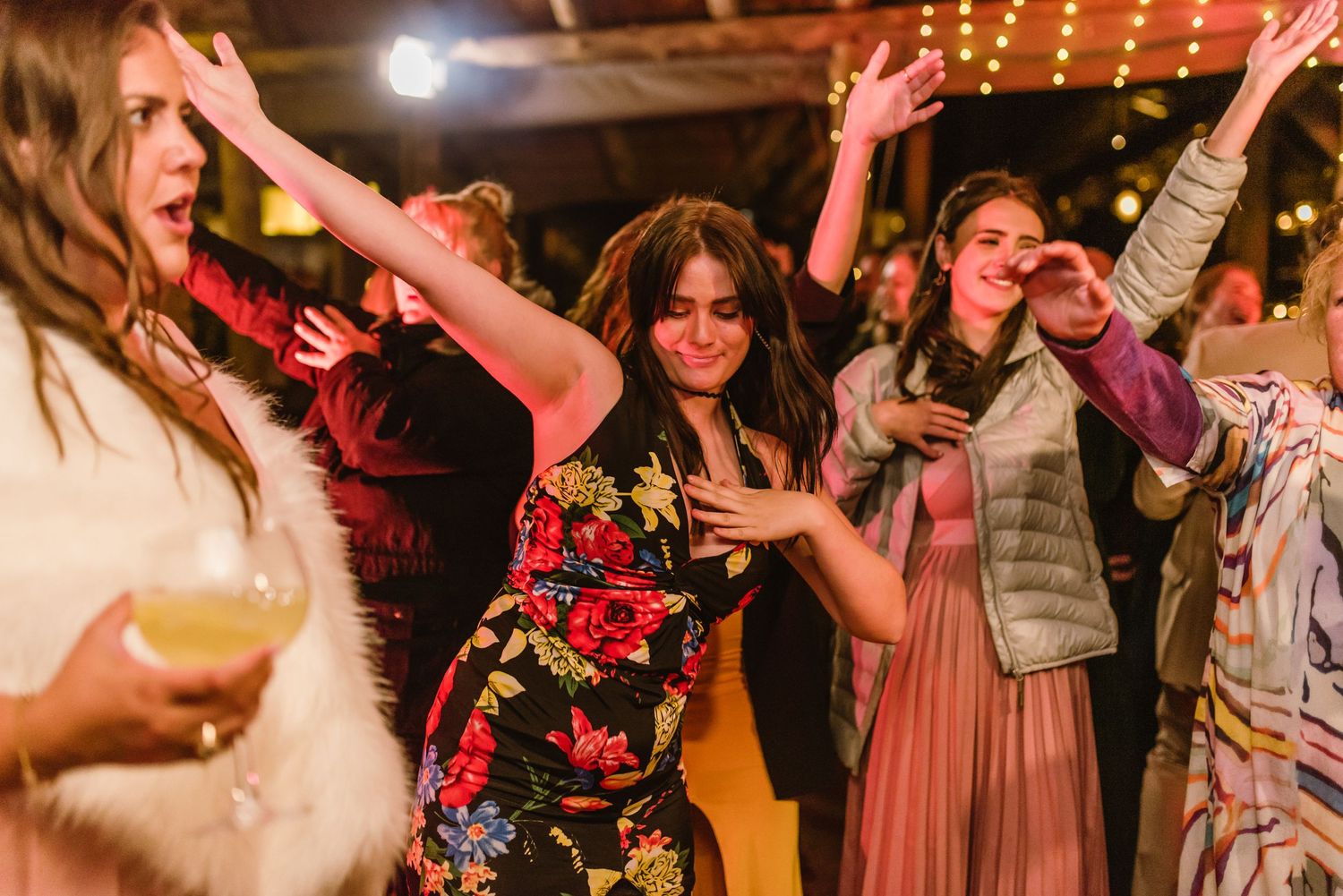 Wedding celebration showing guests dancing with raised arms under string lights at an indoor venue.