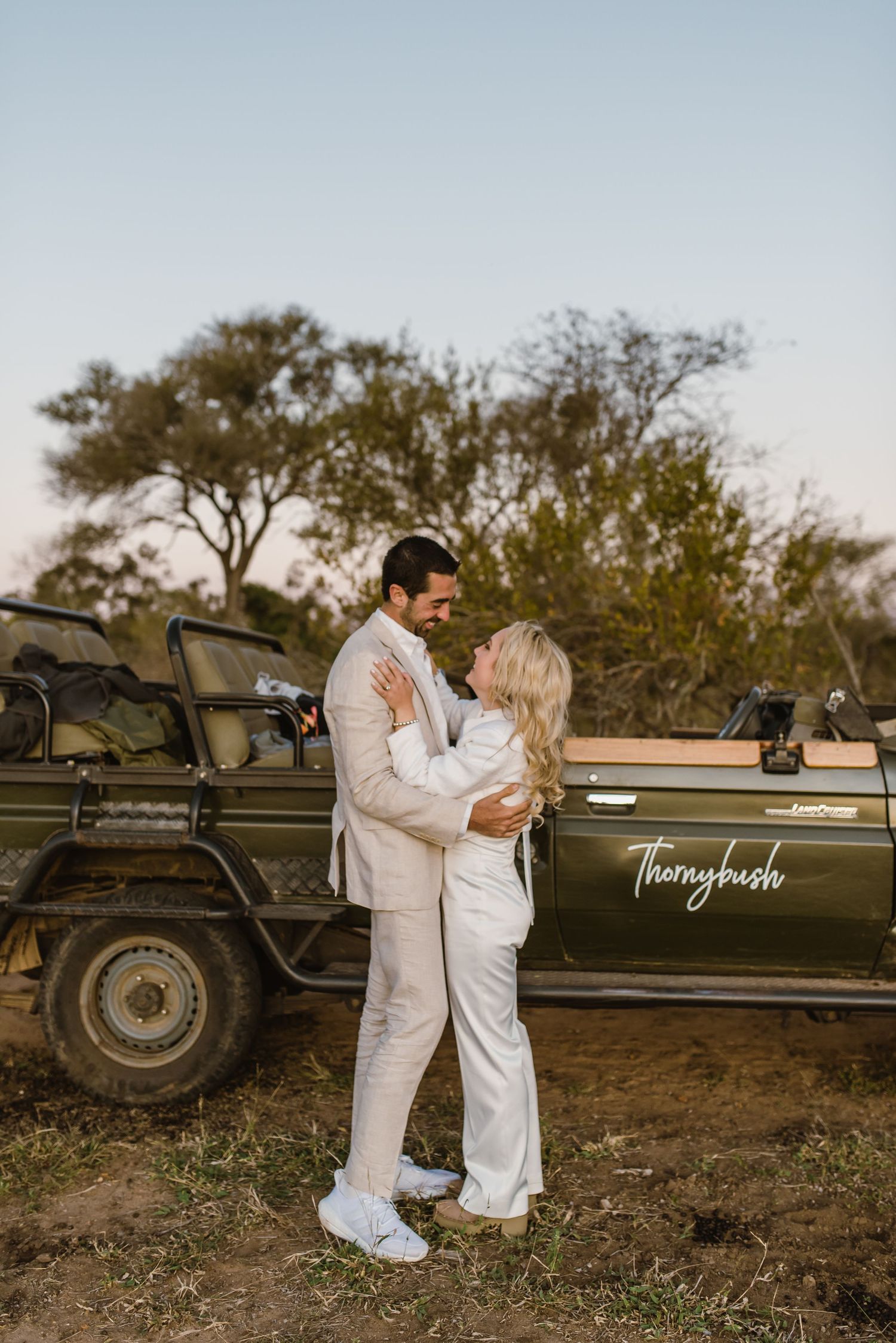 A couple shares romantic moments next to a vintage safari vehicle in a natural outdoor setting.