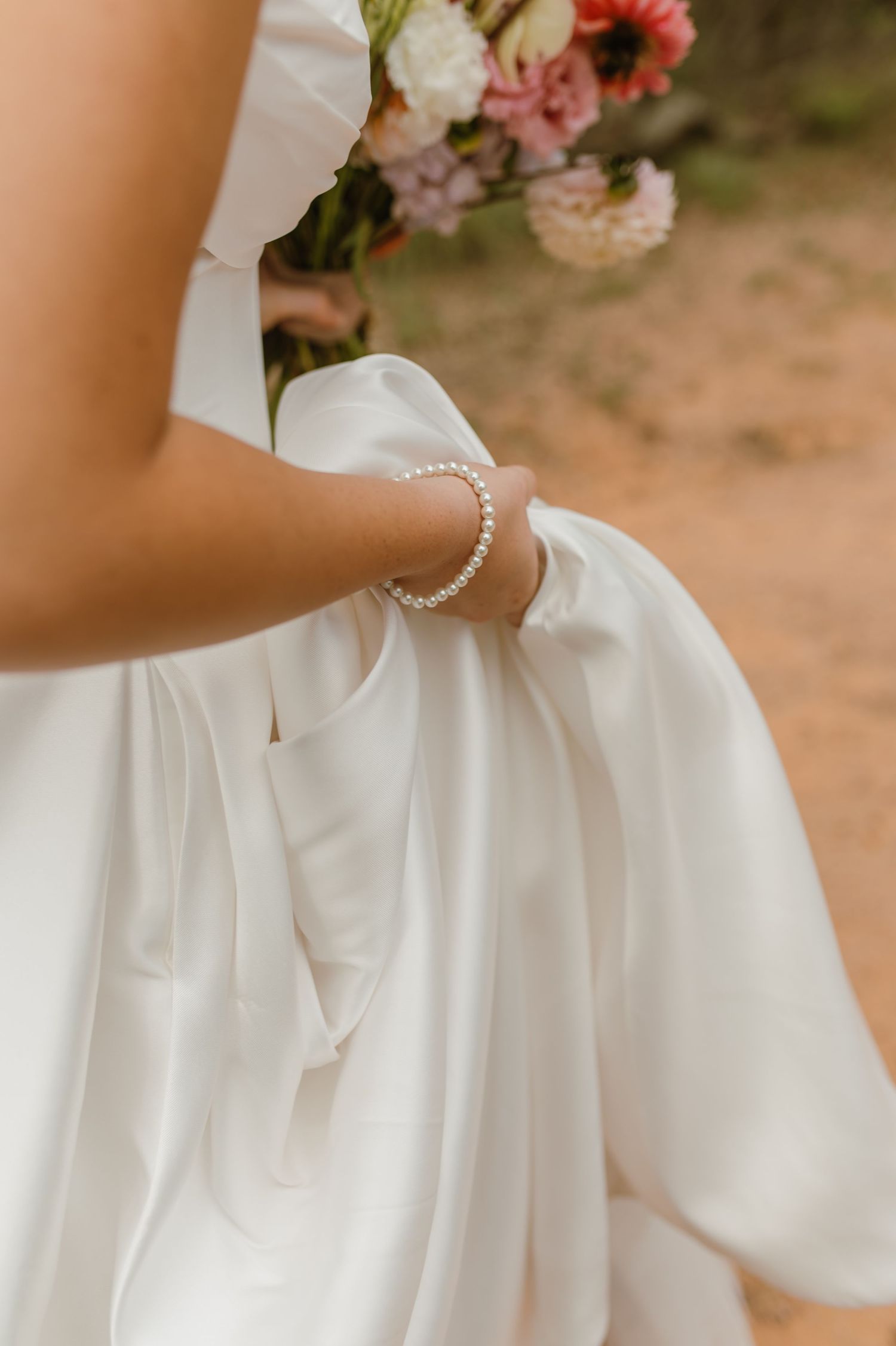 Close-up of bride in white satin gown wearing pearl bracelet while holding colorful wedding bouquet.