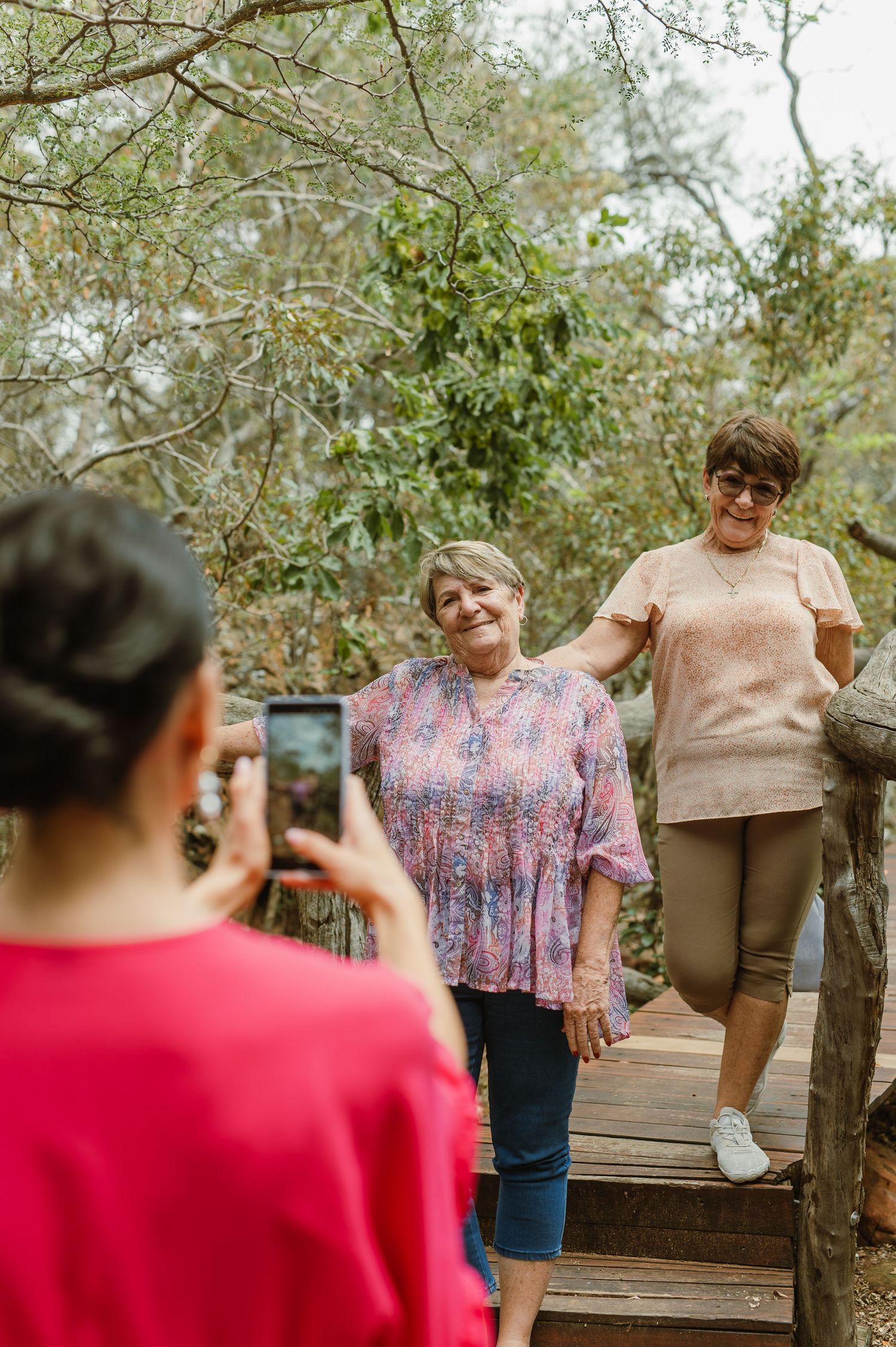 Two older women pose on wooden steps in a forest while a younger woman photographs them with a smartphone.