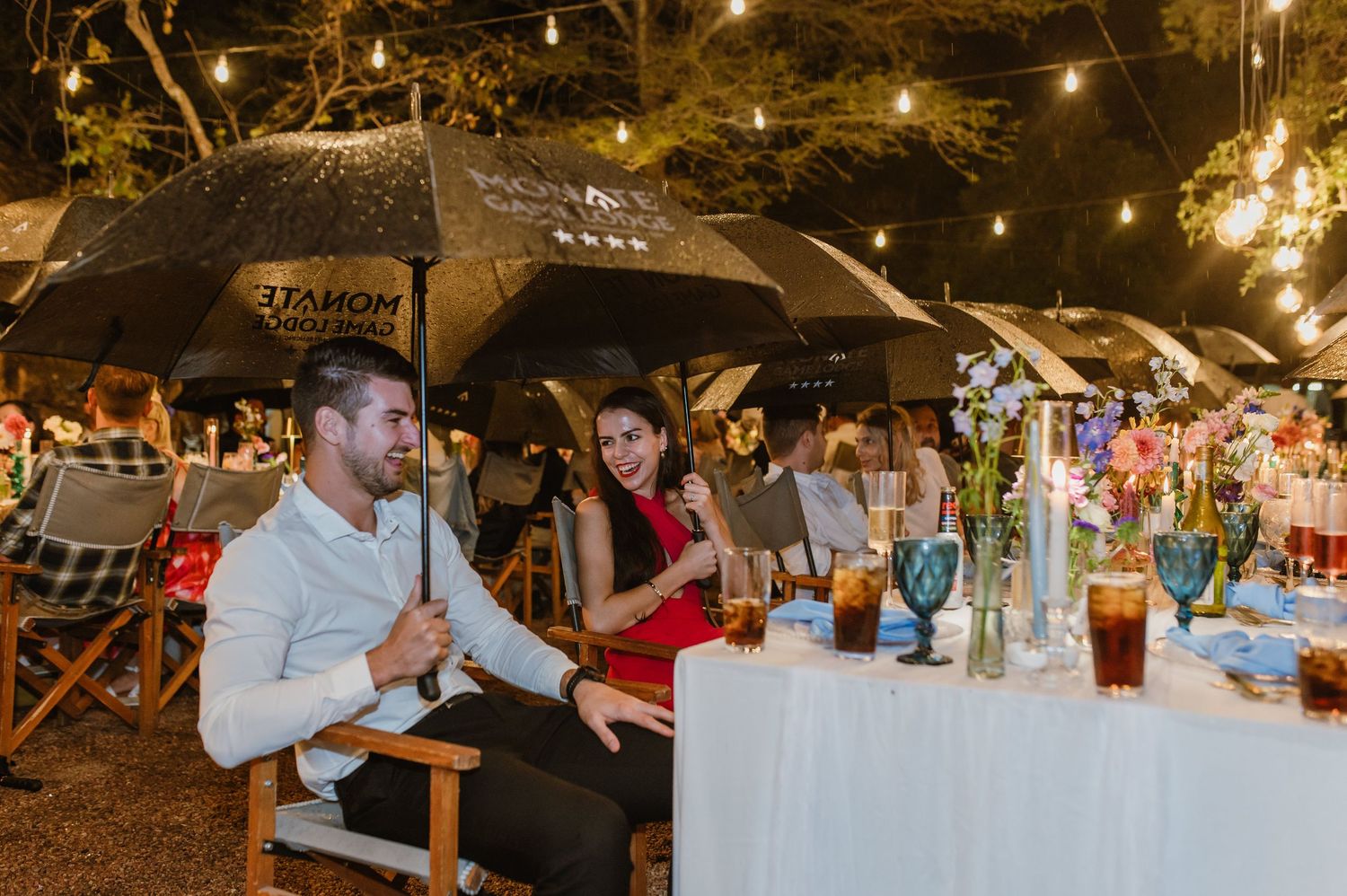 Wedding guests sit under black umbrellas at outdoor evening reception with string lights.