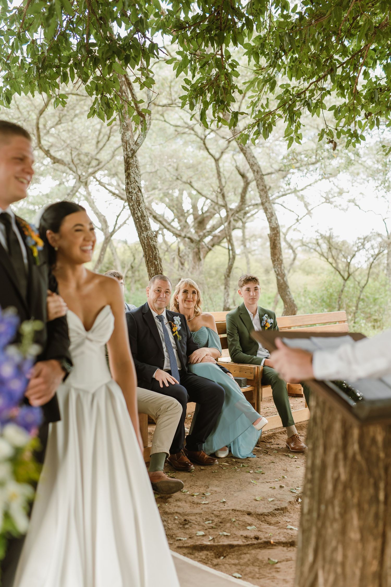Wedding guests smile during outdoor ceremony under trees as bride and groom stand before officiant.