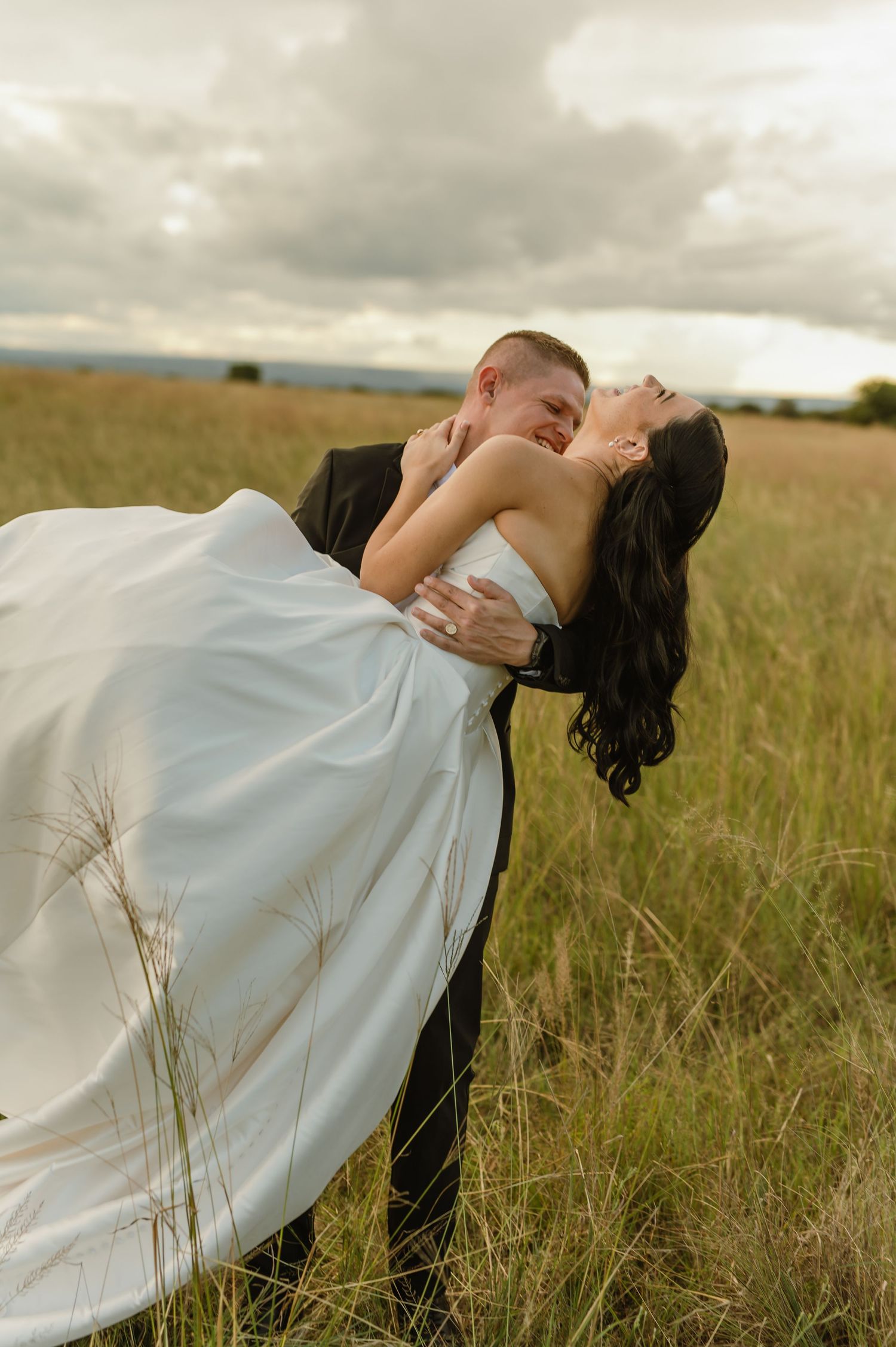 Groom lifting and kissing bride in flowing white gown in golden grass field under cloudy sky.