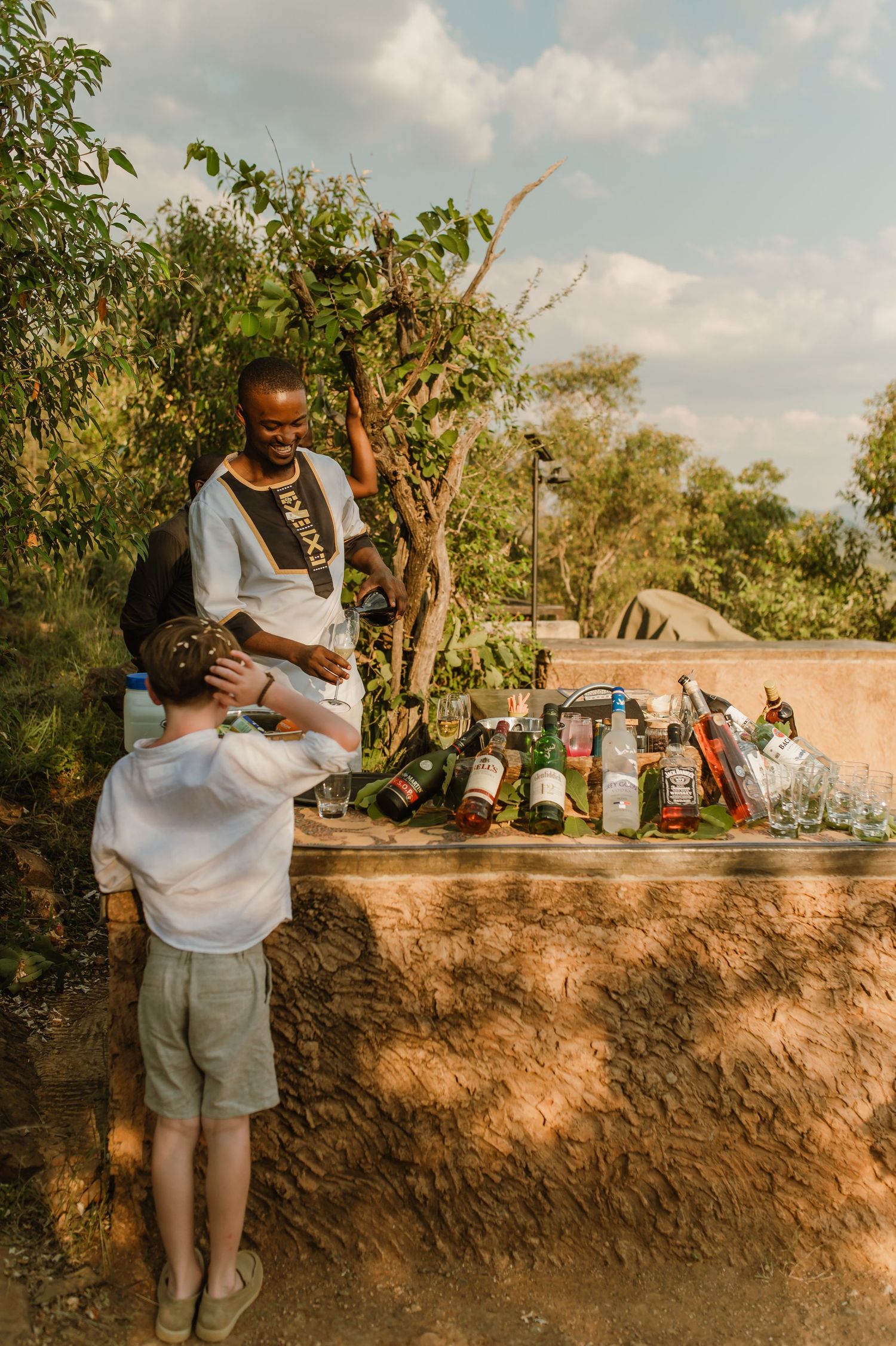 A bartender in traditional African attire serves drinks at an outdoor rustic bar surrounded by lush greenery.
