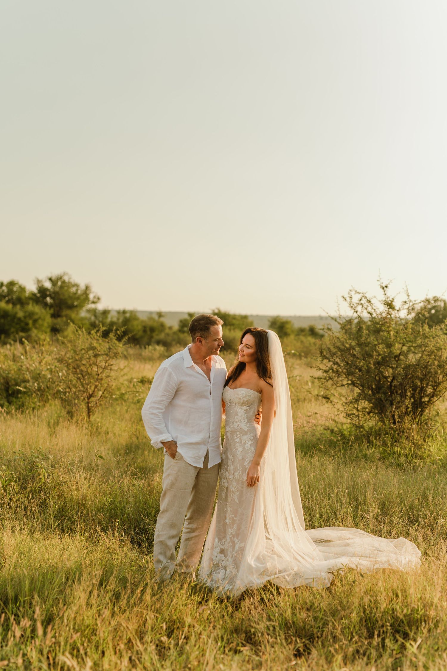Bride and groom share a romantic moment in sun-kissed African savanna, bride wearing lace gown with cathedral veil.
