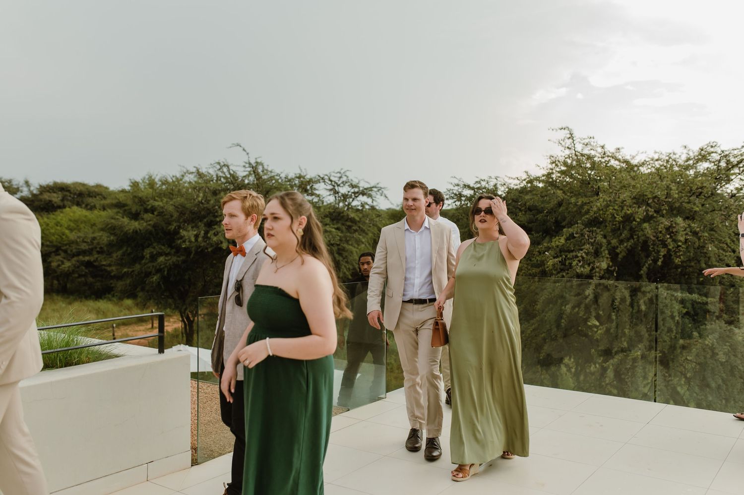 Wedding guests in green dresses and formal attire gather on an outdoor terrace overlooking water at sunset.