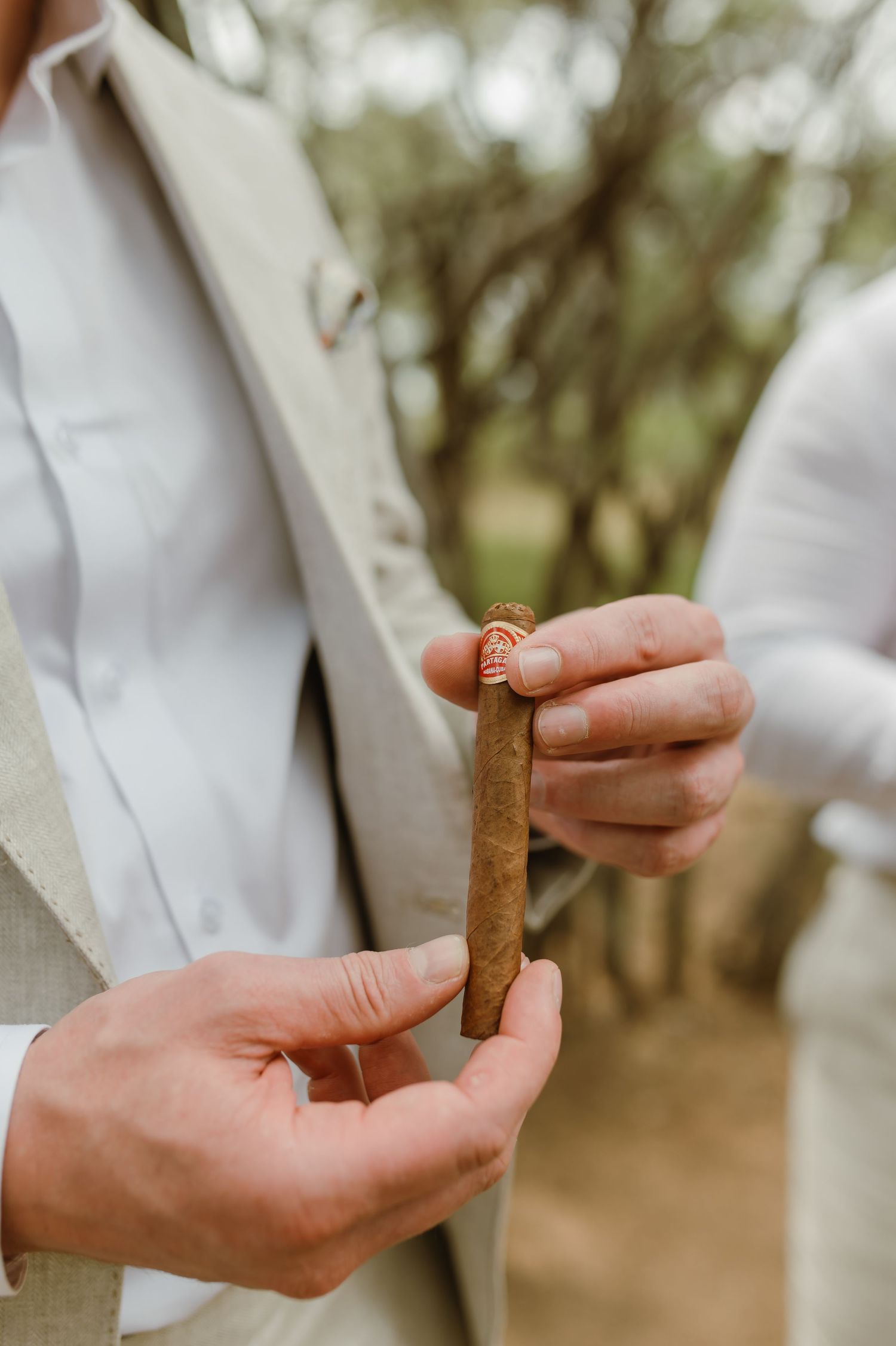 Close-up of hands preparing a cigar outdoors in white formal attire.