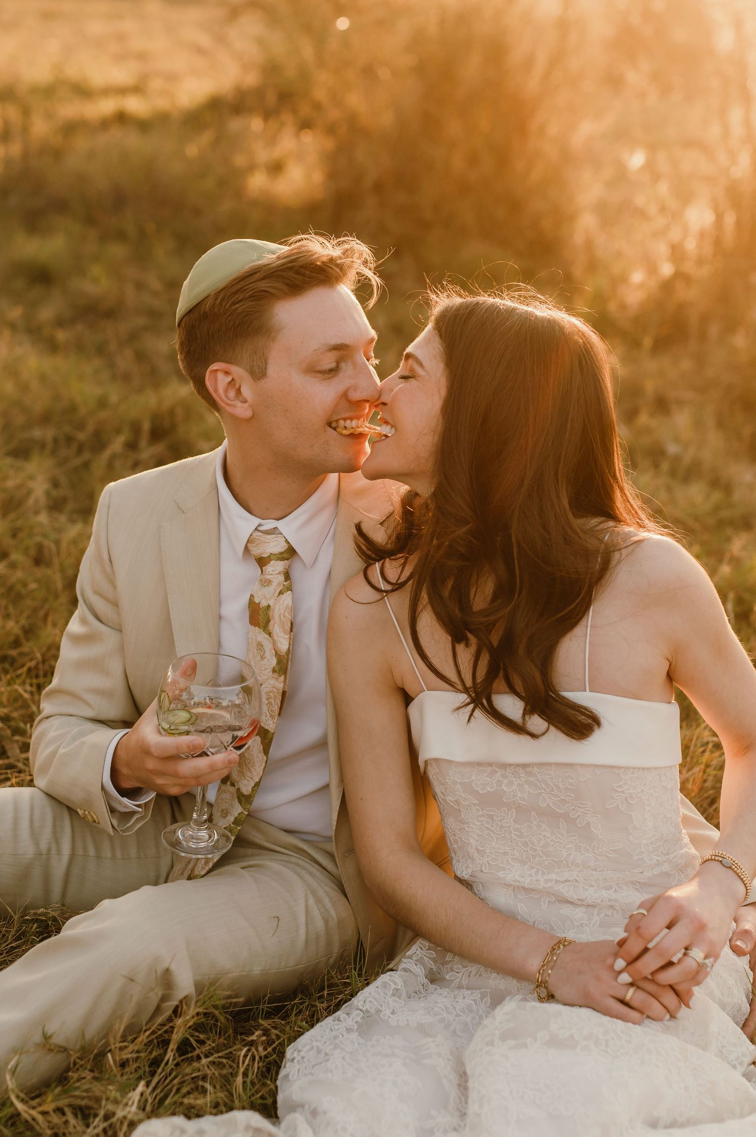 A couple in wedding attire shares a romantic kiss at golden hour, sitting in a sunlit field with champagne glasses.