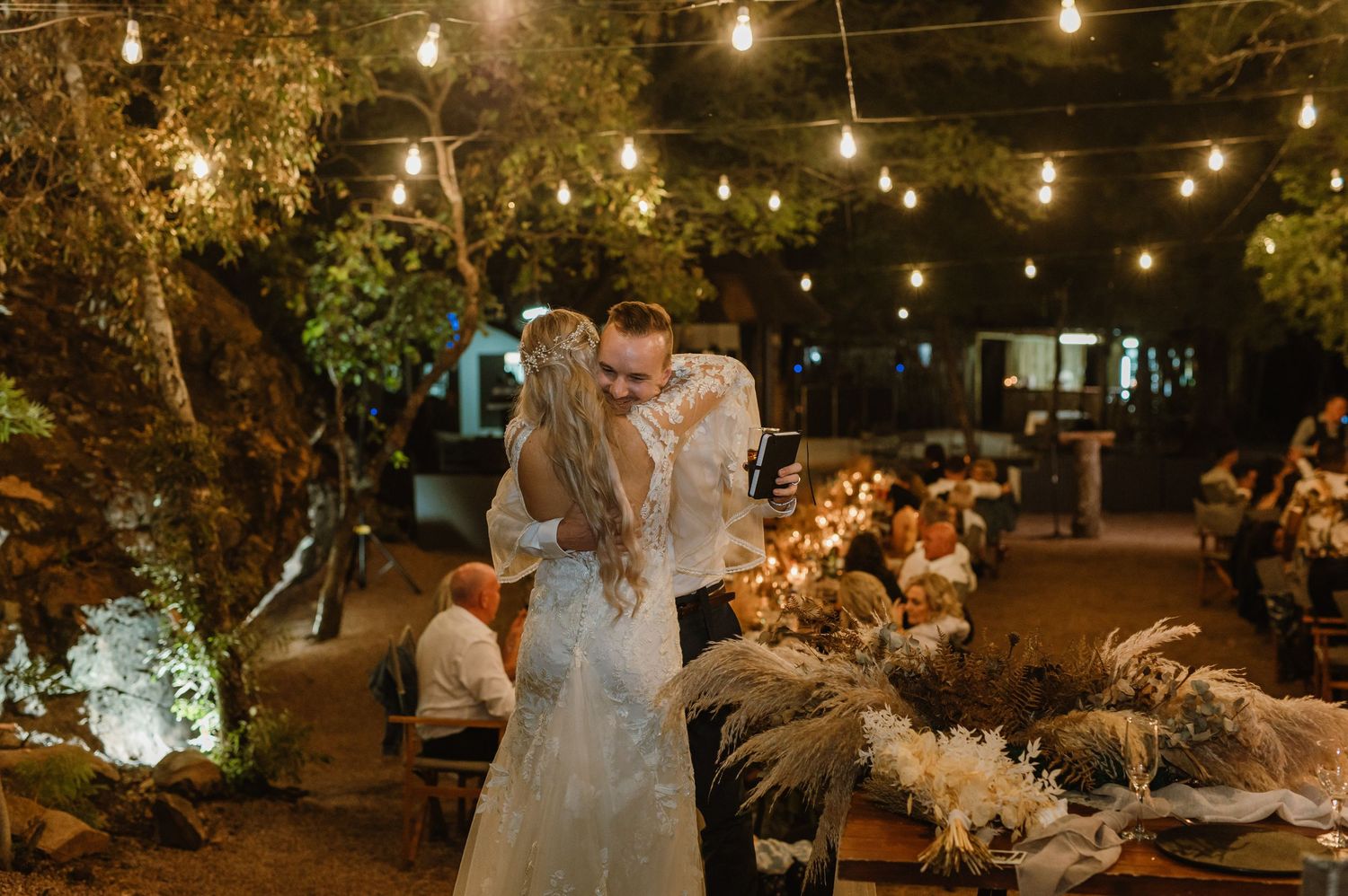 Wedding couple dancing under string lights at outdoor evening reception with guests watching.