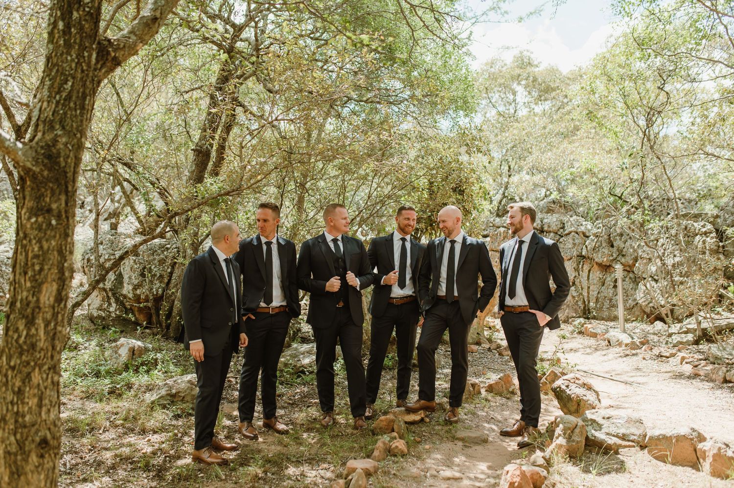 Groomsmen in black suits lined up outdoors under tree canopy.