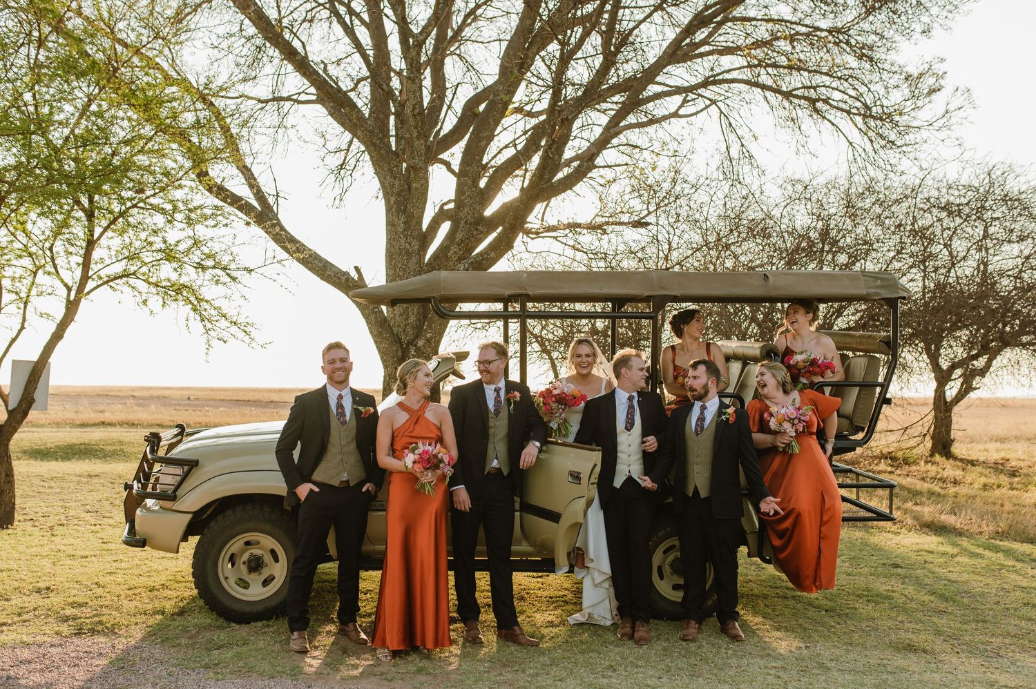 Wedding group photo with bridesmaids in rust-colored dresses posed by vintage car at sunset.