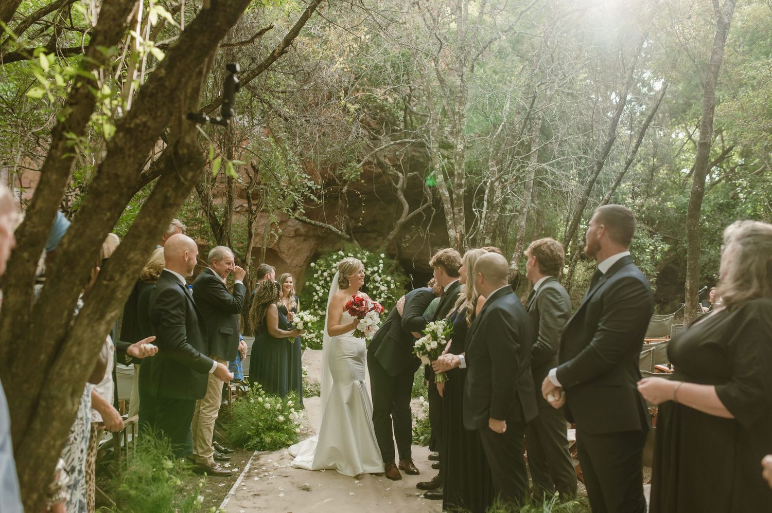 Wedding ceremony taking place on a scenic wooded path with guests gathered under a natural canopy of trees.