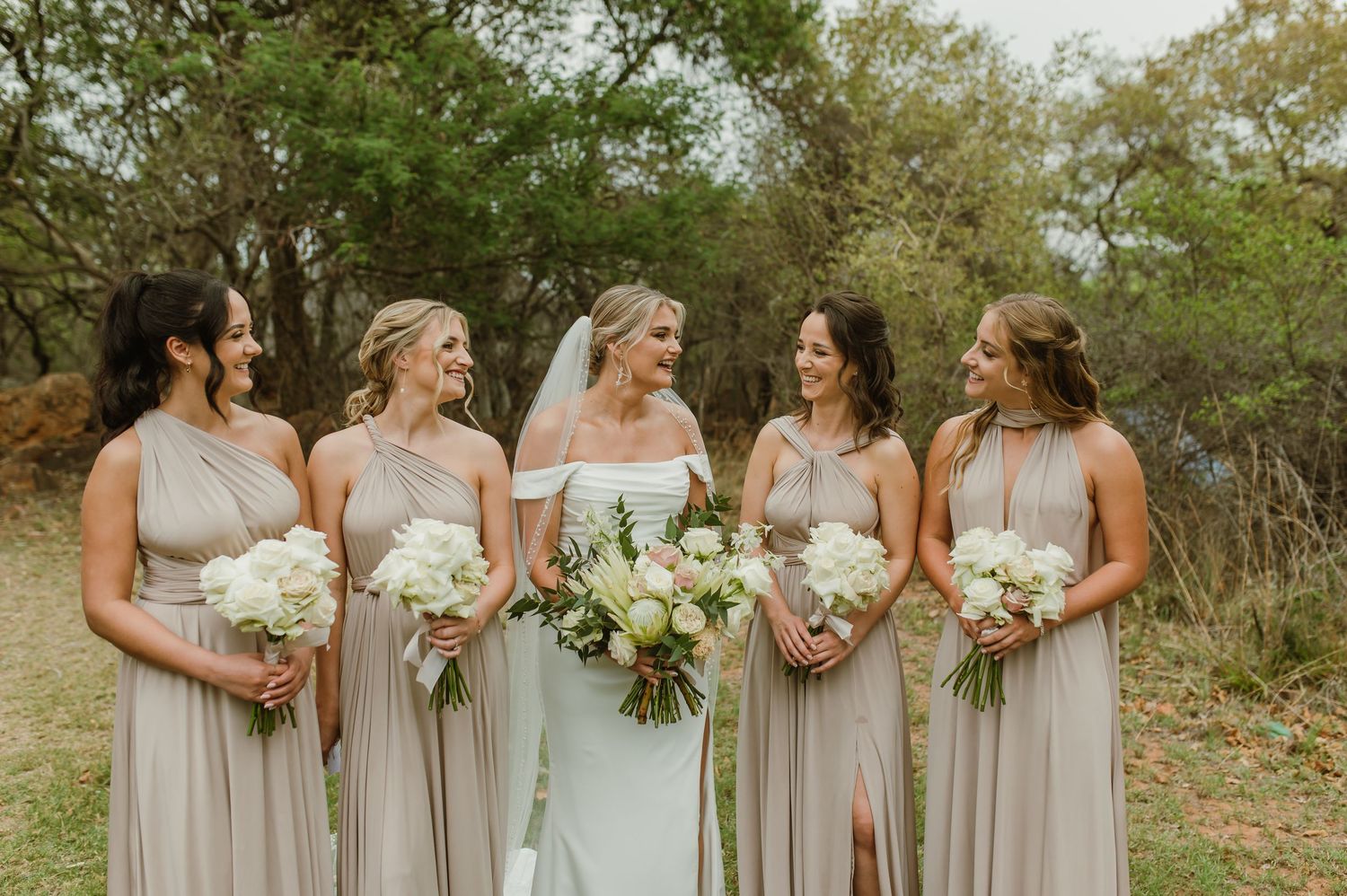 Wedding party in neutral dresses poses together holding bouquets in outdoor garden setting.