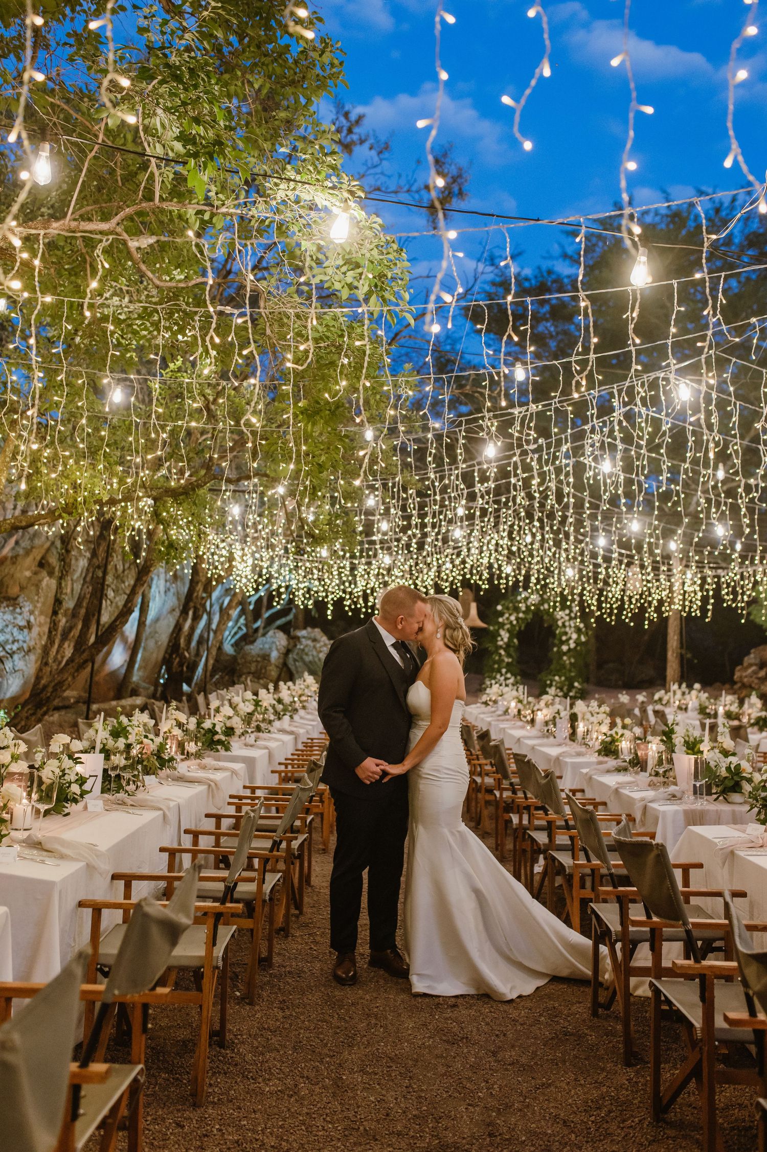 Elegant outdoor wedding venue with rows of wooden chairs under twinkling string lights.