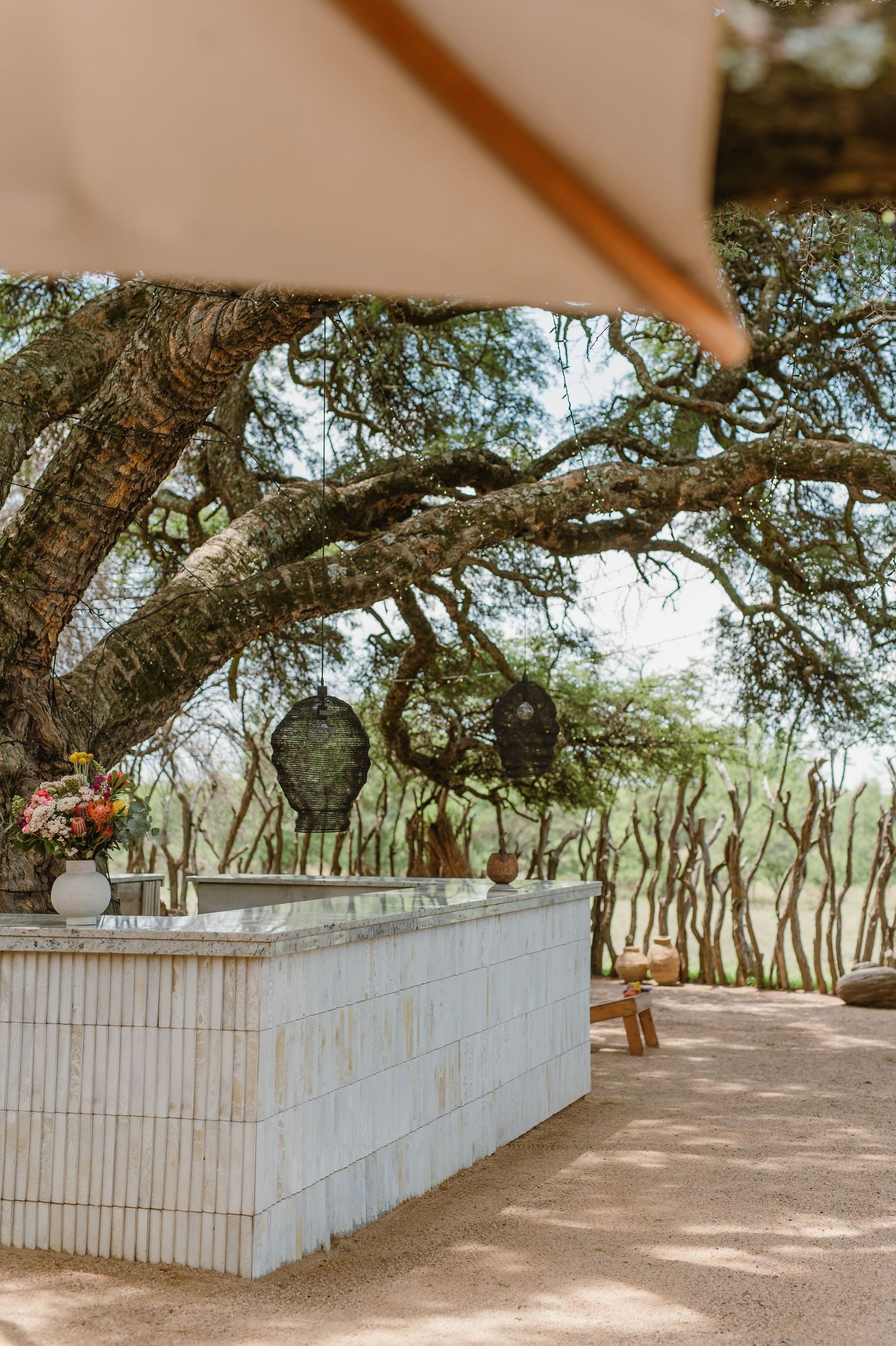 Outdoor bar setup with white brick counter and hanging lanterns under natural tree coverage.