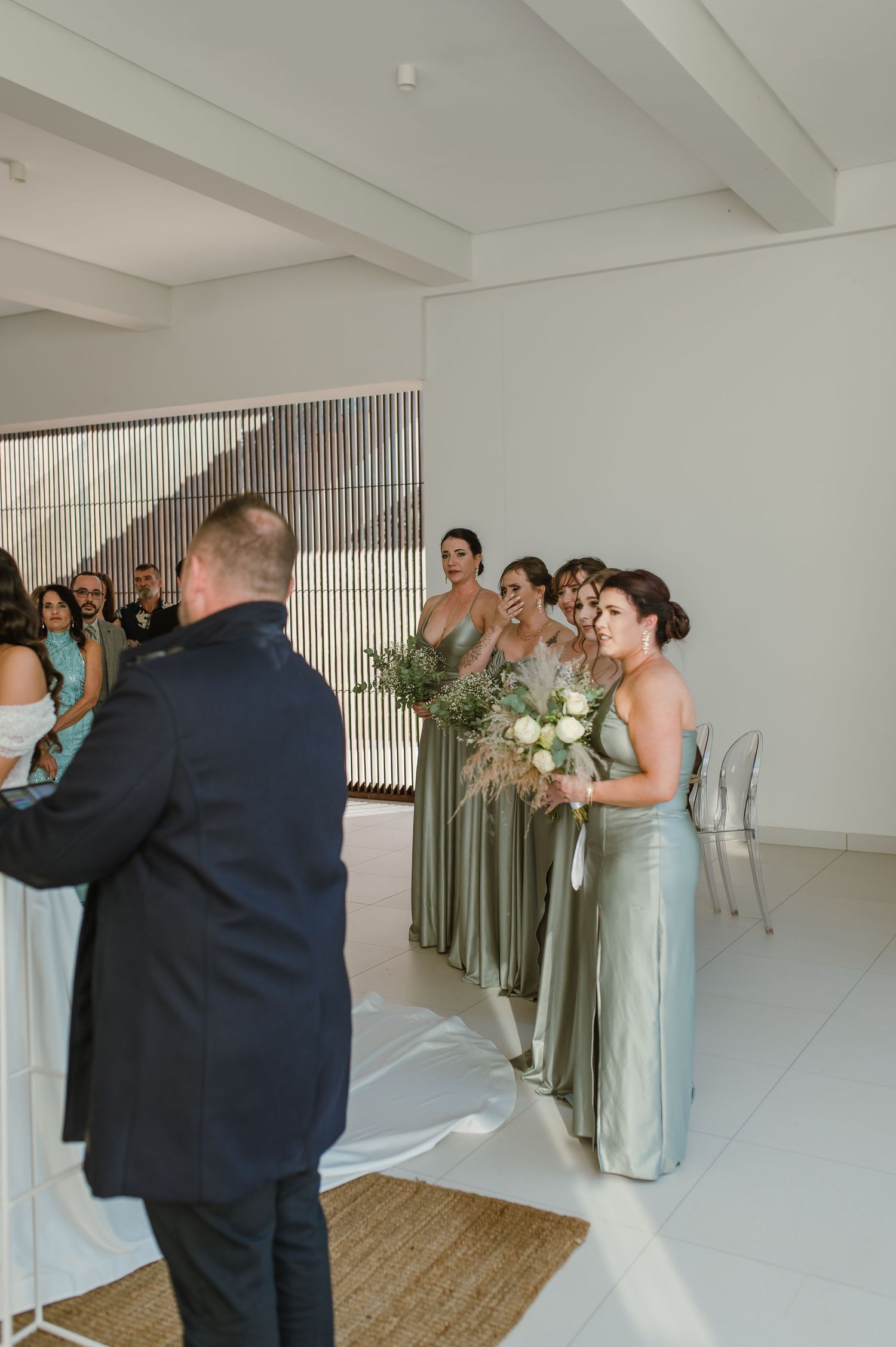 Bridesmaids in sage green dresses line up against a white wall during a wedding ceremony.