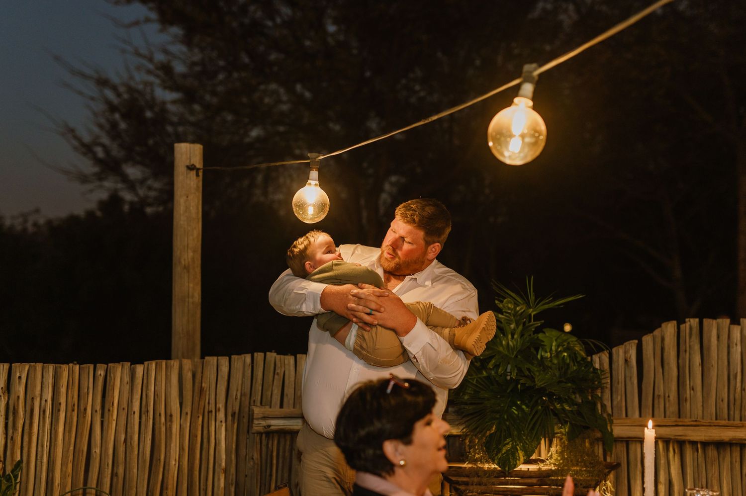 String lights illuminate the evening sky at an outdoor wedding venue with wooden fencing.