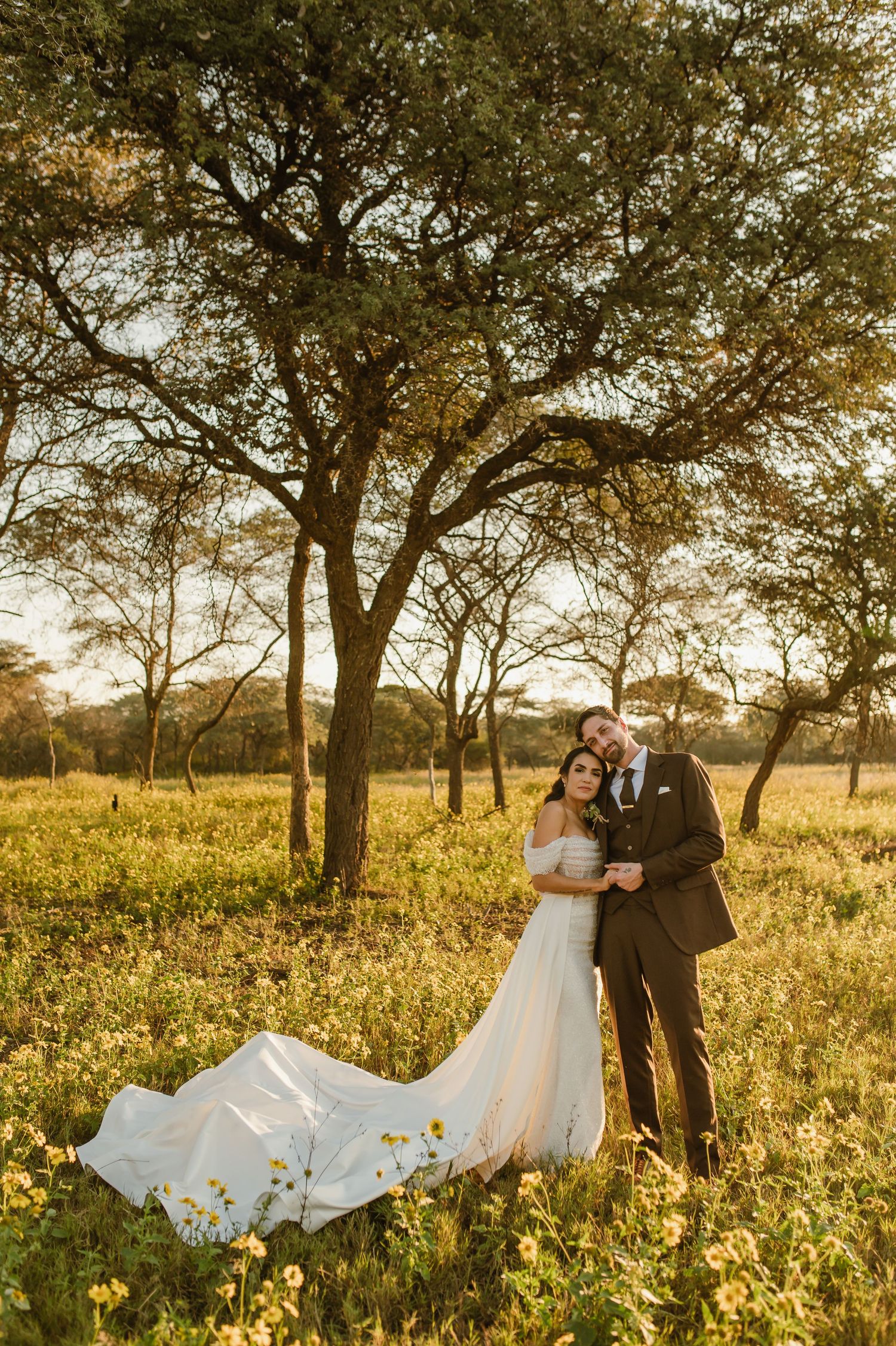 A wedding couple poses in a wildflower field with dramatic tree silhouettes during sunset.