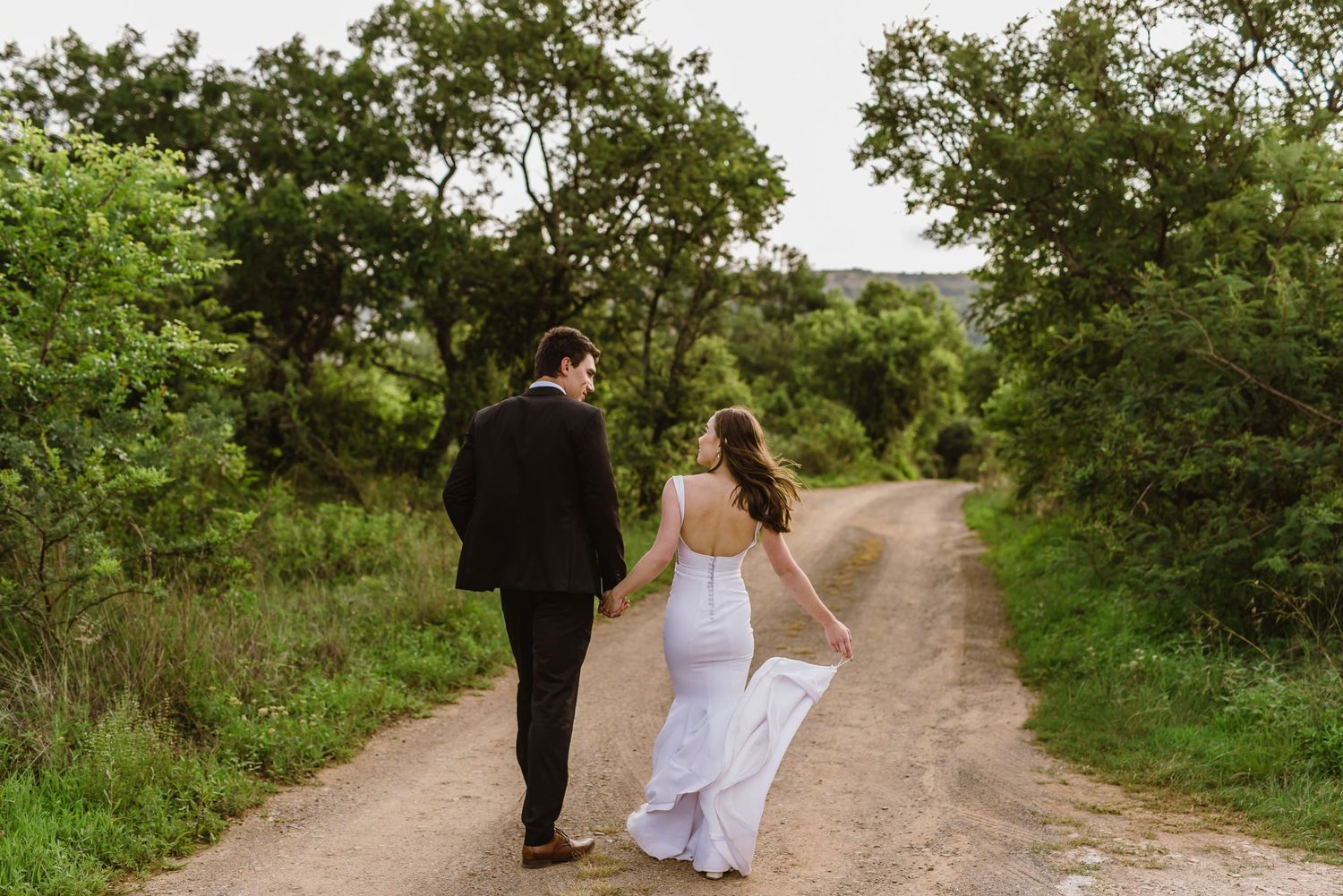 A couple walks hand in hand down a winding dirt path surrounded by lush vegetation.