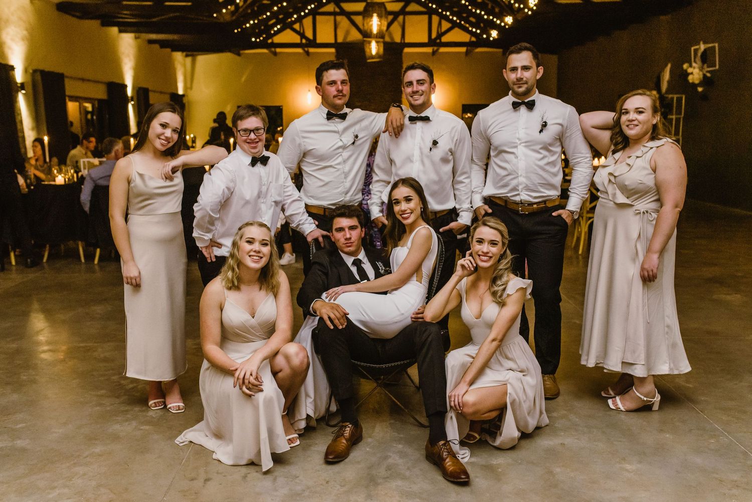 Wedding party poses together in formal attire at an indoor reception venue.