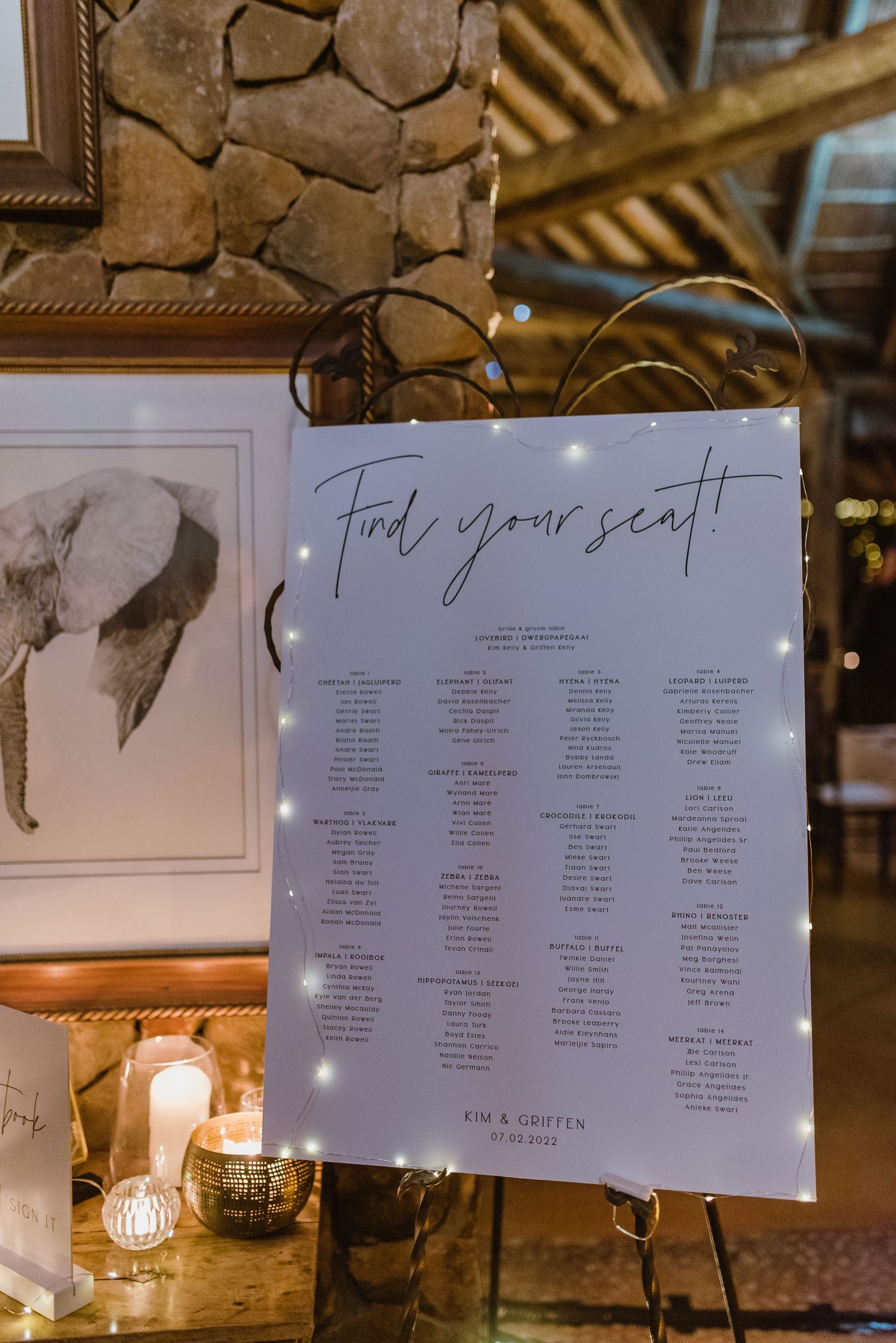 Illuminated seating chart display with multiple paper signs hanging from rustic wooden beams.