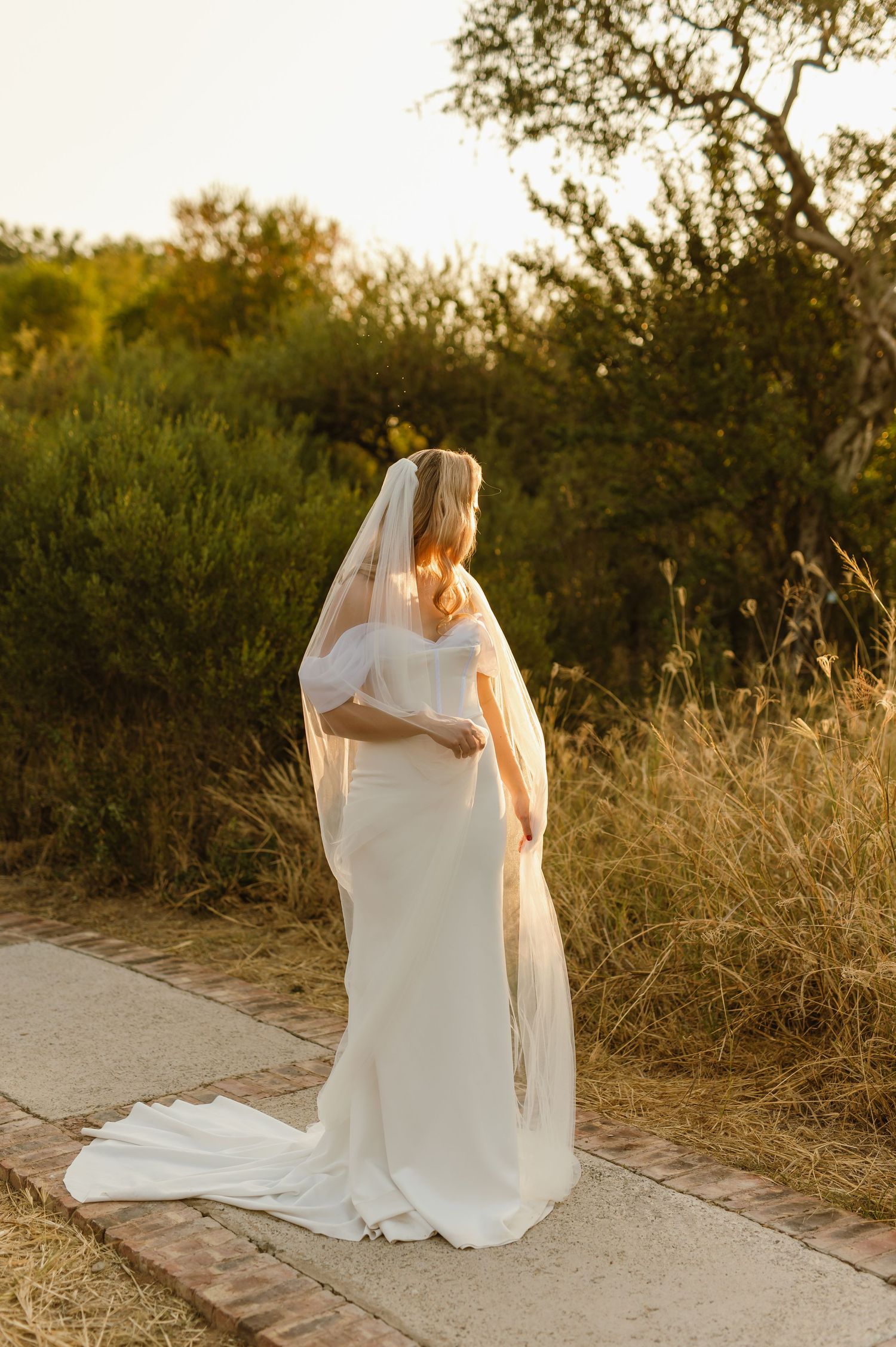 Bride in flowing white dress sits peacefully on a desert path at sunset.