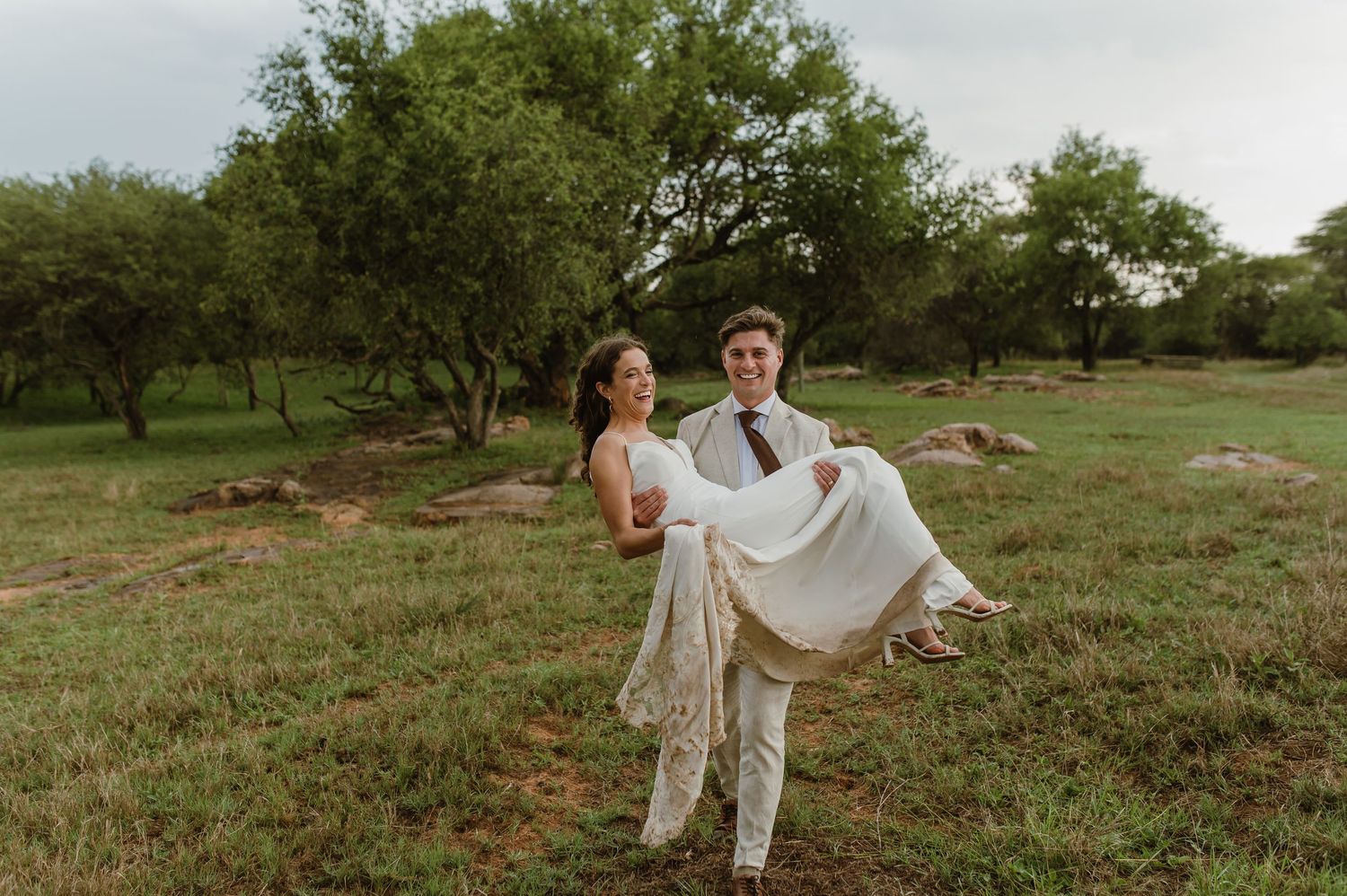 A wedding couple shares a joyful moment as the groom carries the bride in a rustic outdoor setting.