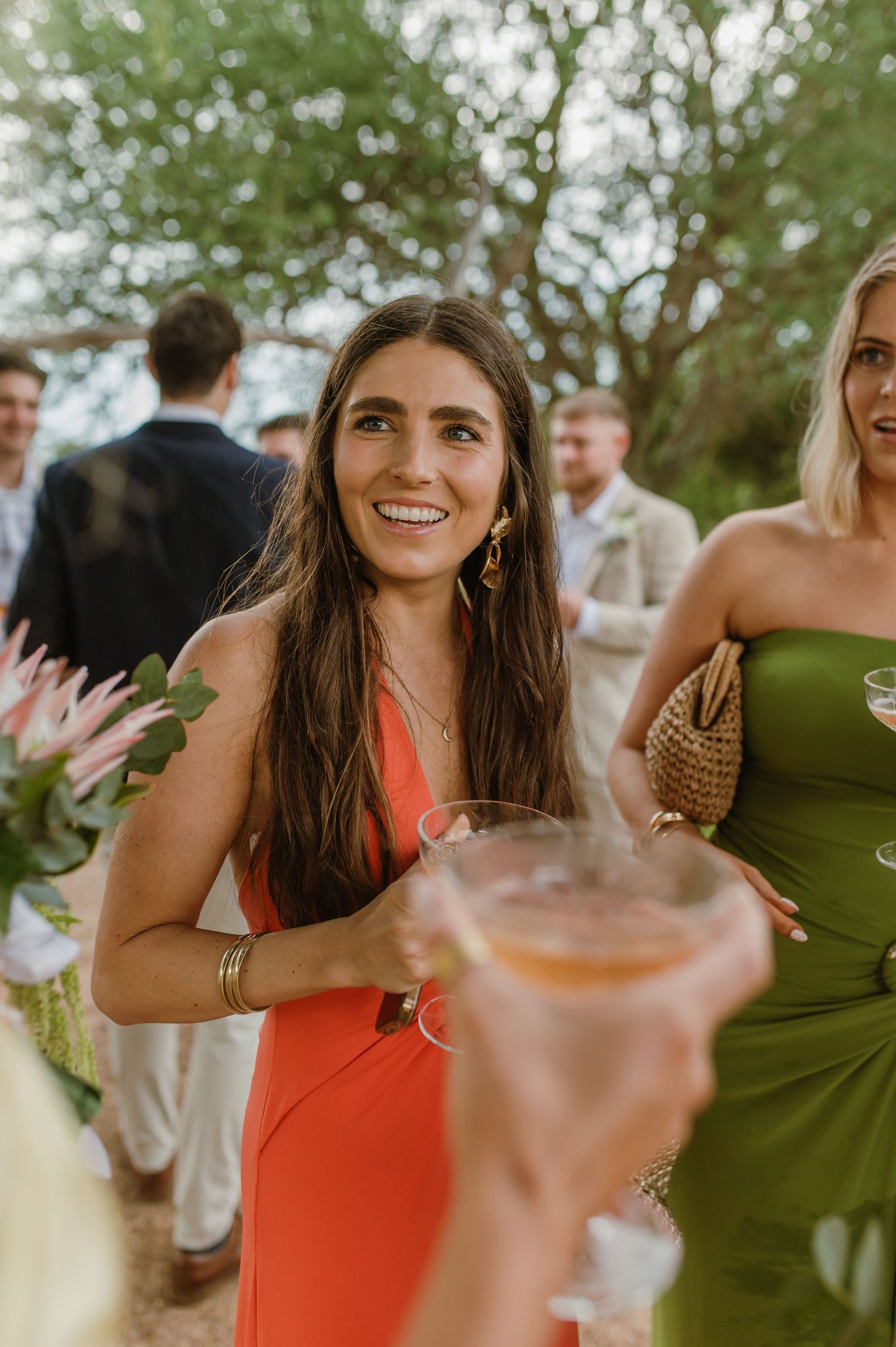 Wedding attendees in bright dresses celebrate with drinks under trees.