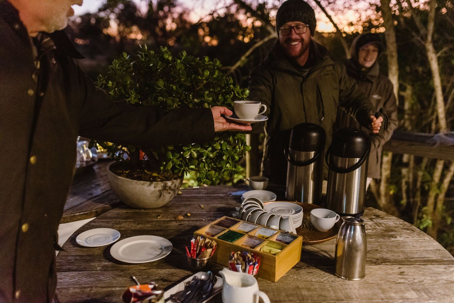 People enjoying coffee and snacks at a rustic outdoor wooden table during sunset with camping gear and supplies.