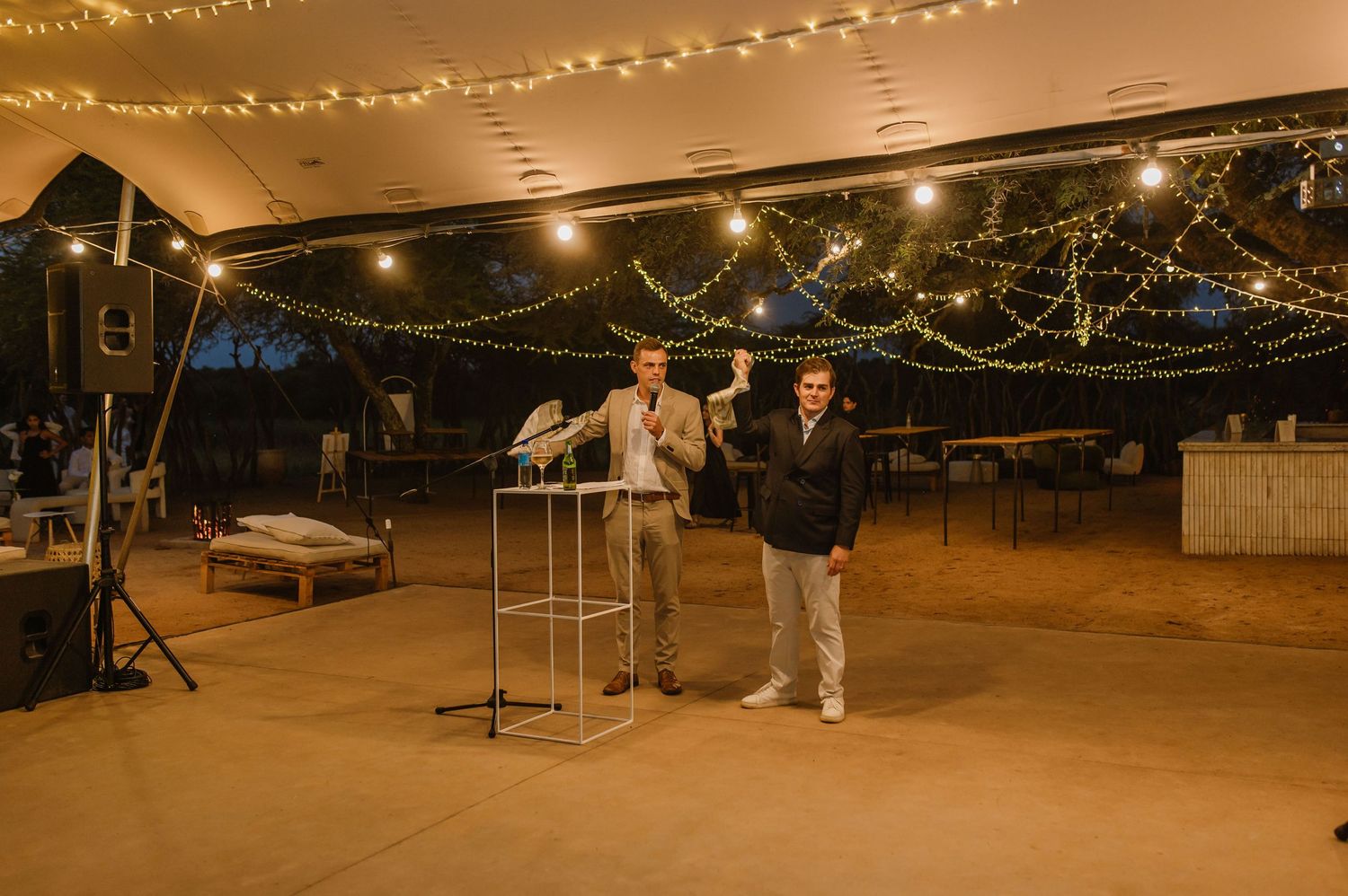 Two people speaking at microphones under string lights at an outdoor evening event.