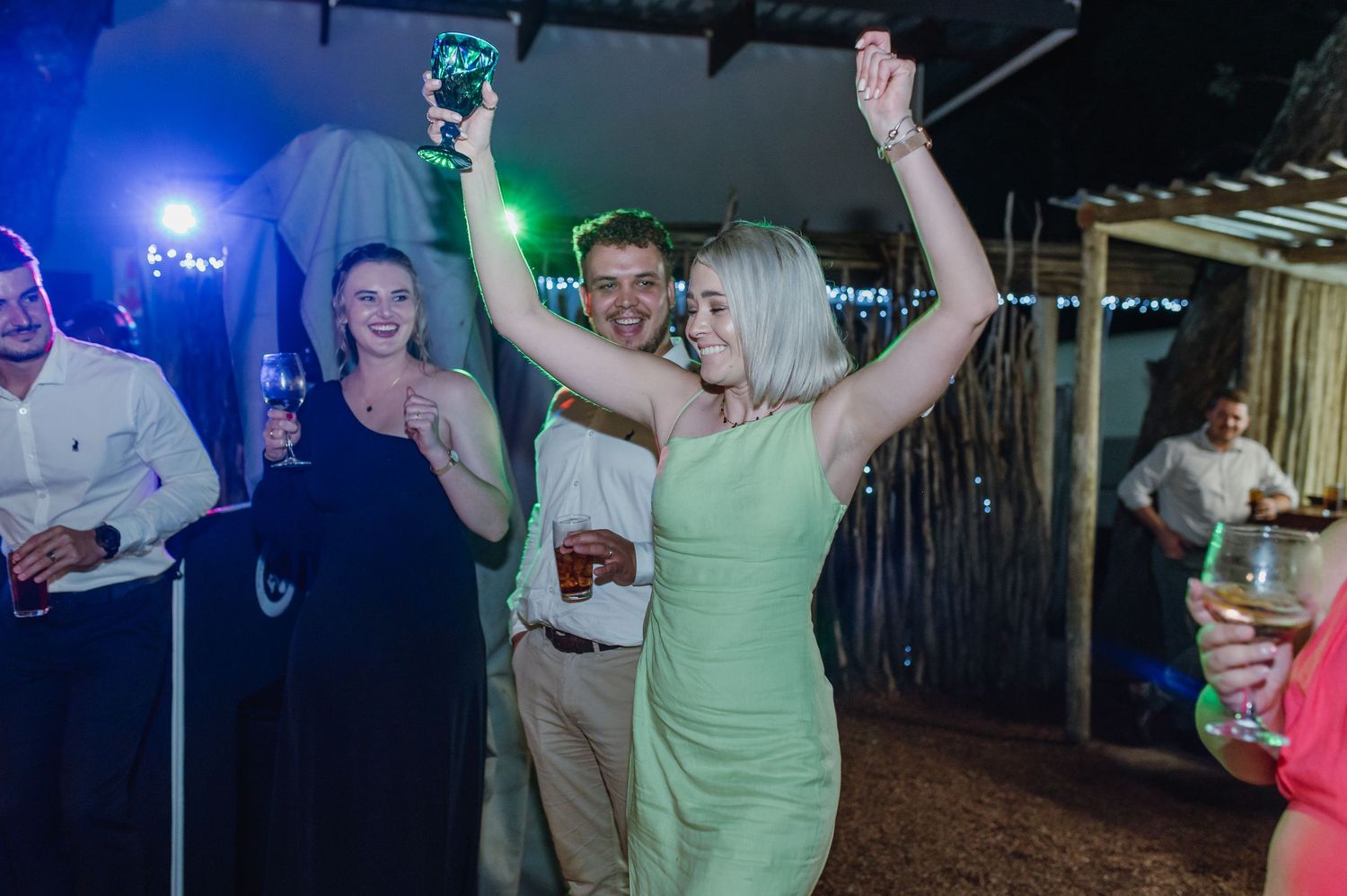 Wedding guest in green dress dances joyfully with arms raised at lively reception party.