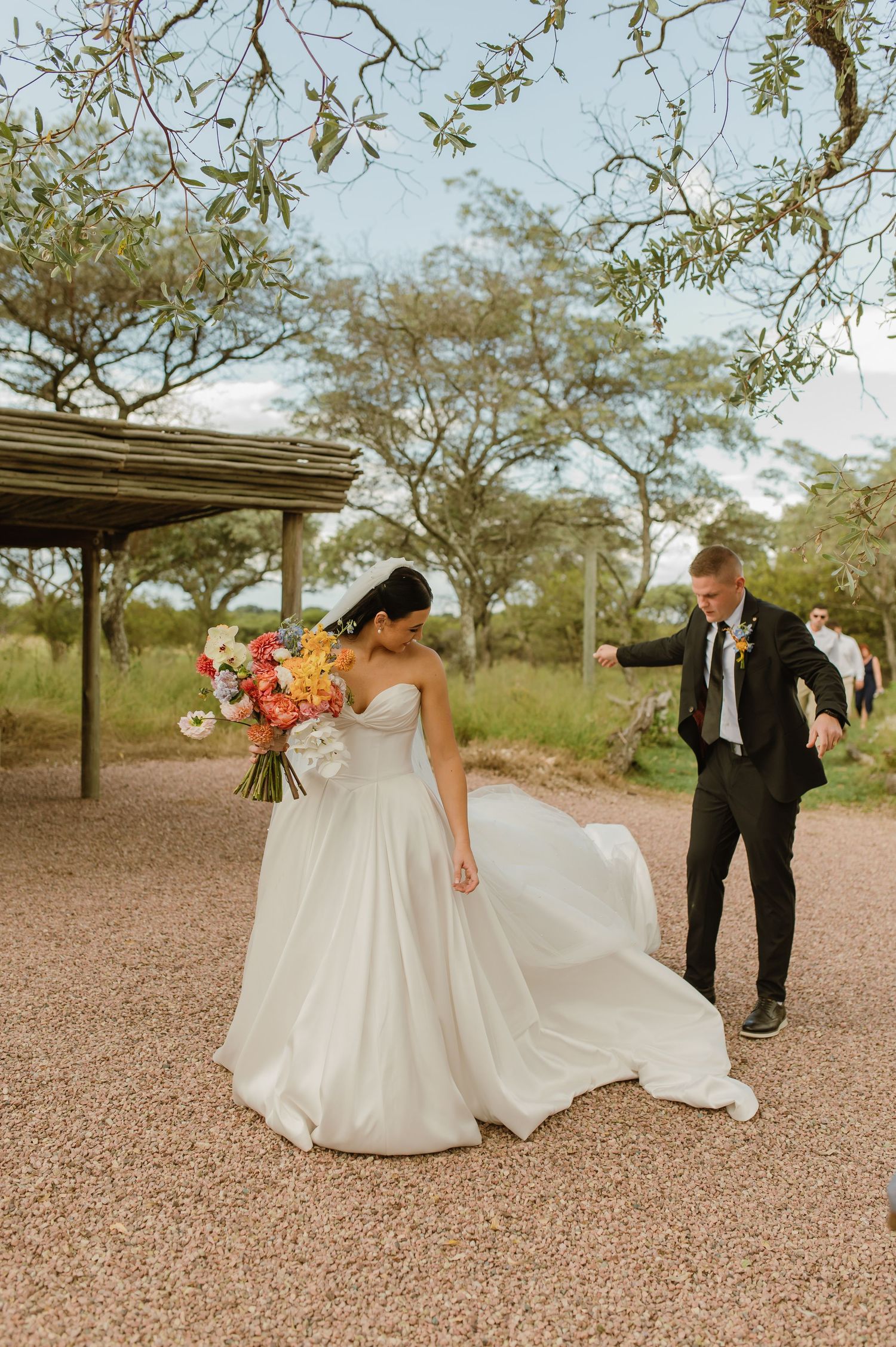 Bride in strapless white ball gown with colorful bouquet as groom in black suit adjusts her dress train.