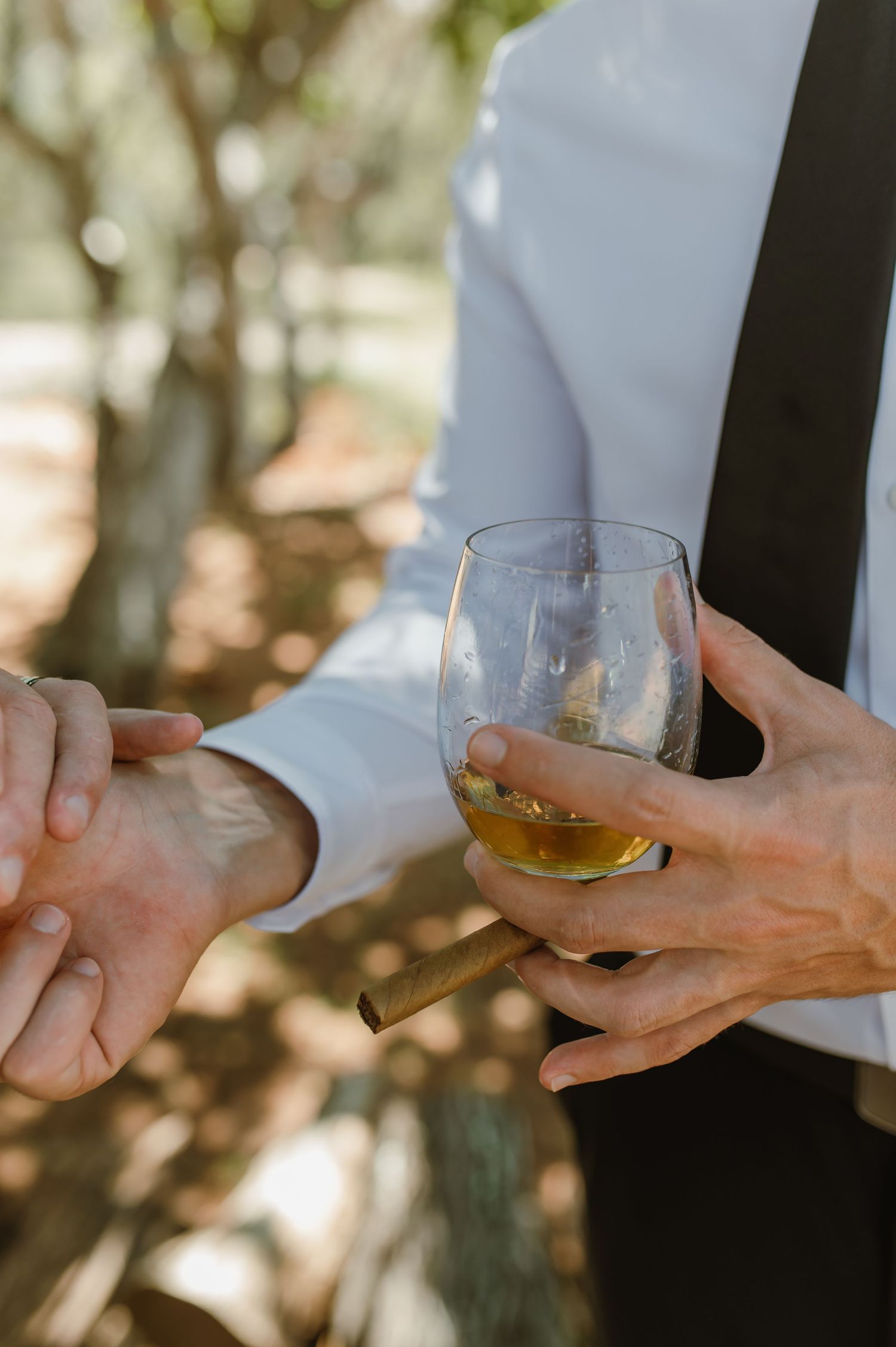 Groom holding whiskey glass and cigar while shaking hands outdoors at wedding celebration.