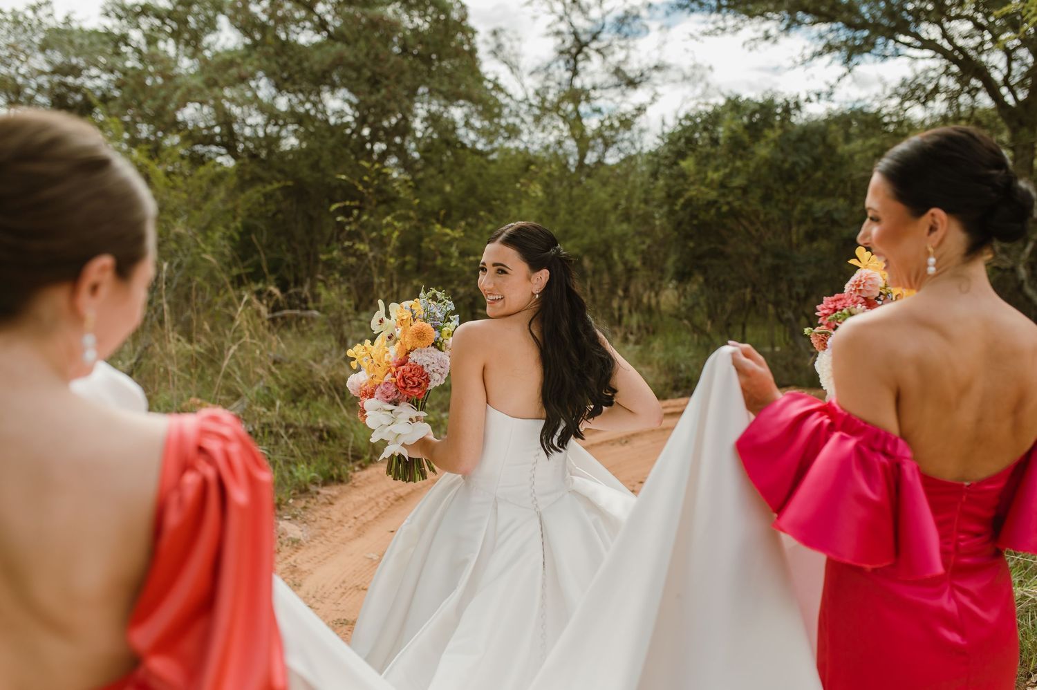 Bride in strapless white gown holding colorful bouquet while bridesmaids in coral and pink dresses assist on dirt path.