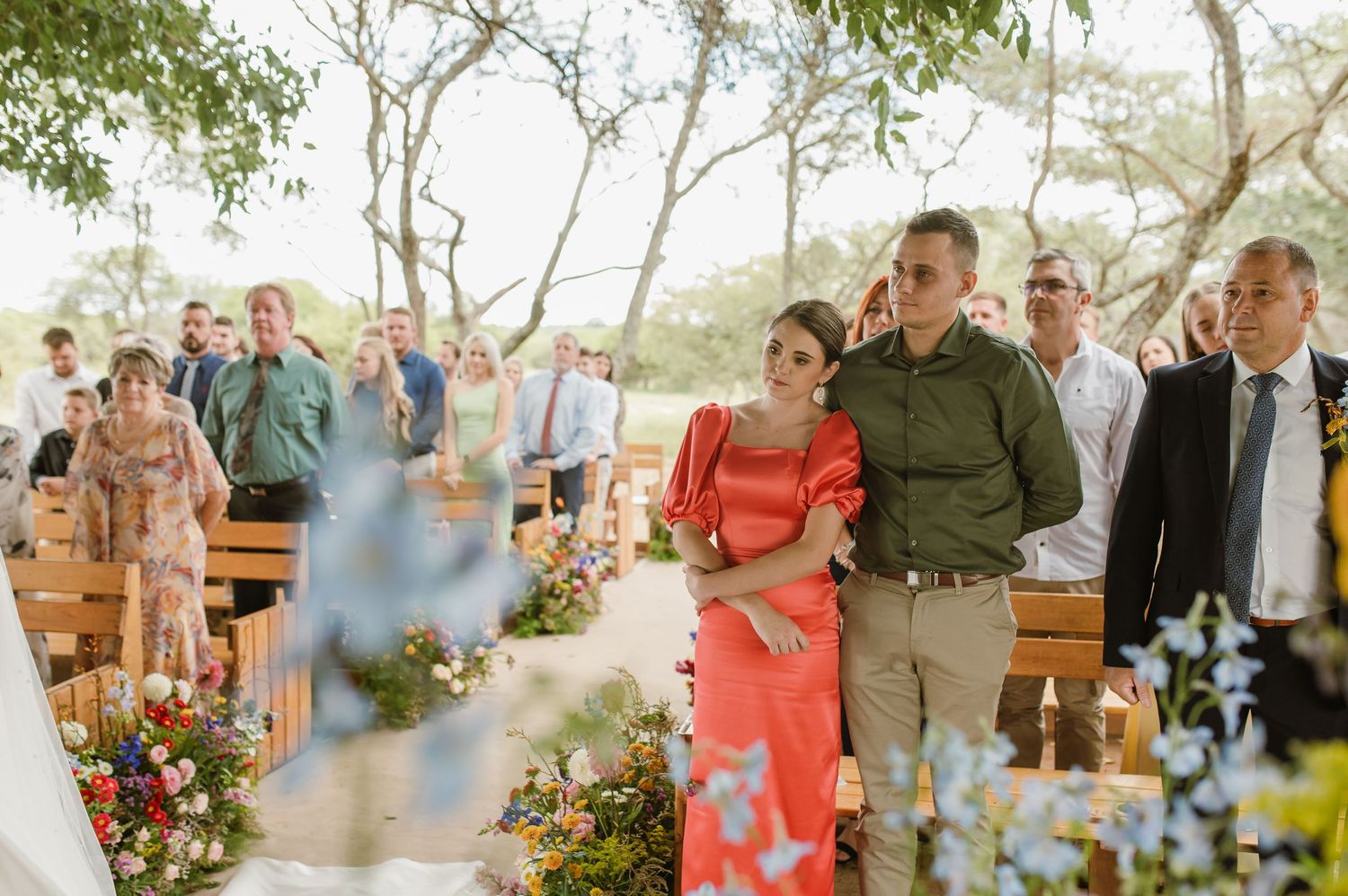 Couple embracing in red and green outfits watching outdoor wedding ceremony among seated guests.