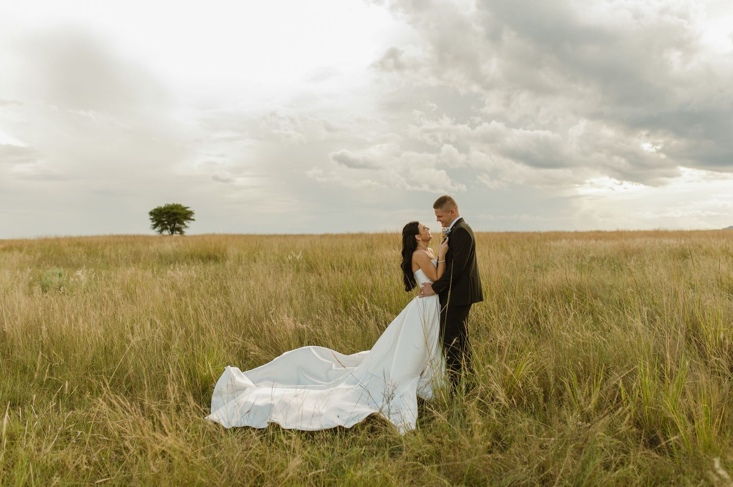 Bride and groom embrace romantically in vast golden grass field under dramatic cloudy sky at sunset.