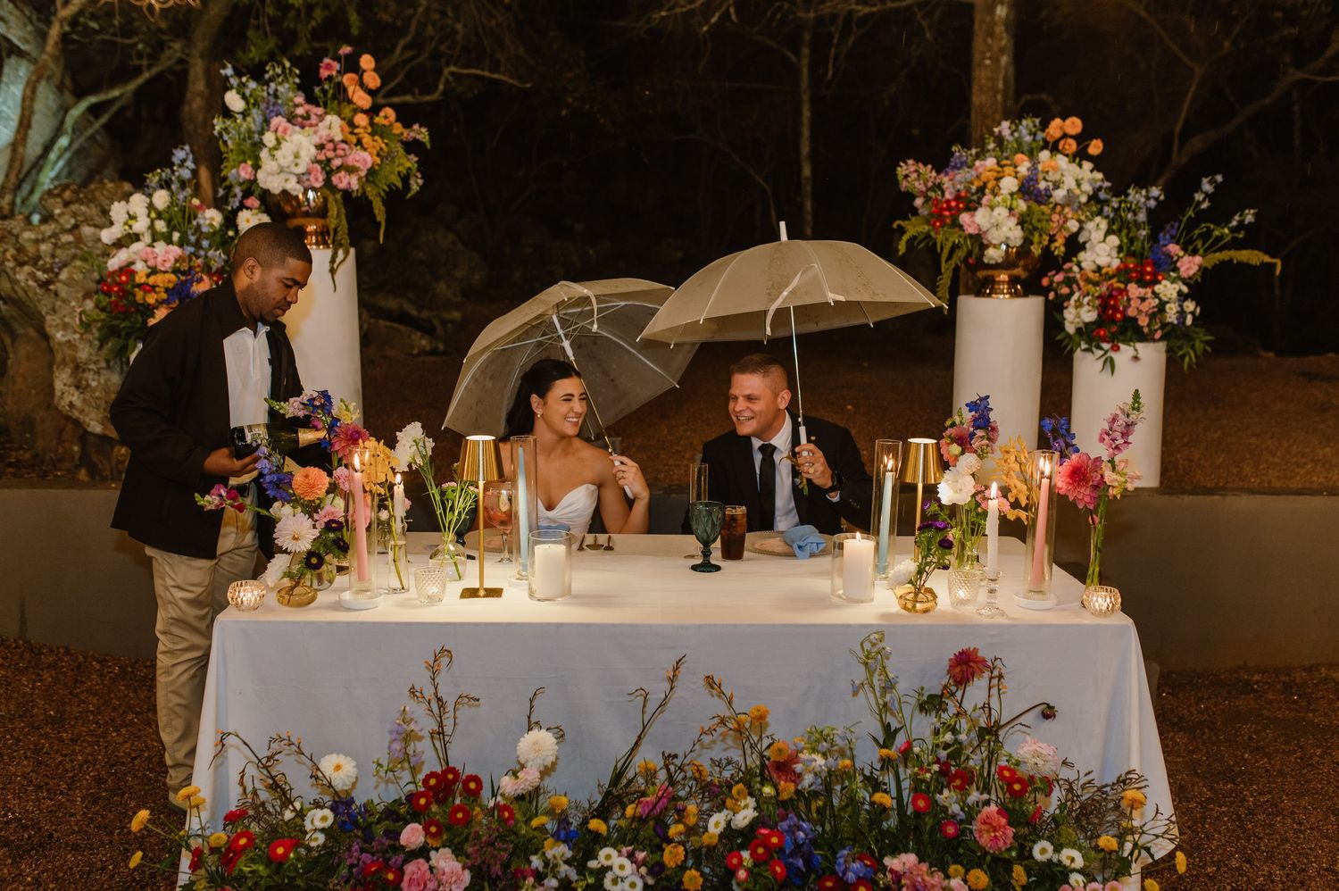 Bride and groom seated at sweetheart table under umbrellas surrounded by vibrant wildflower arrangements at night.