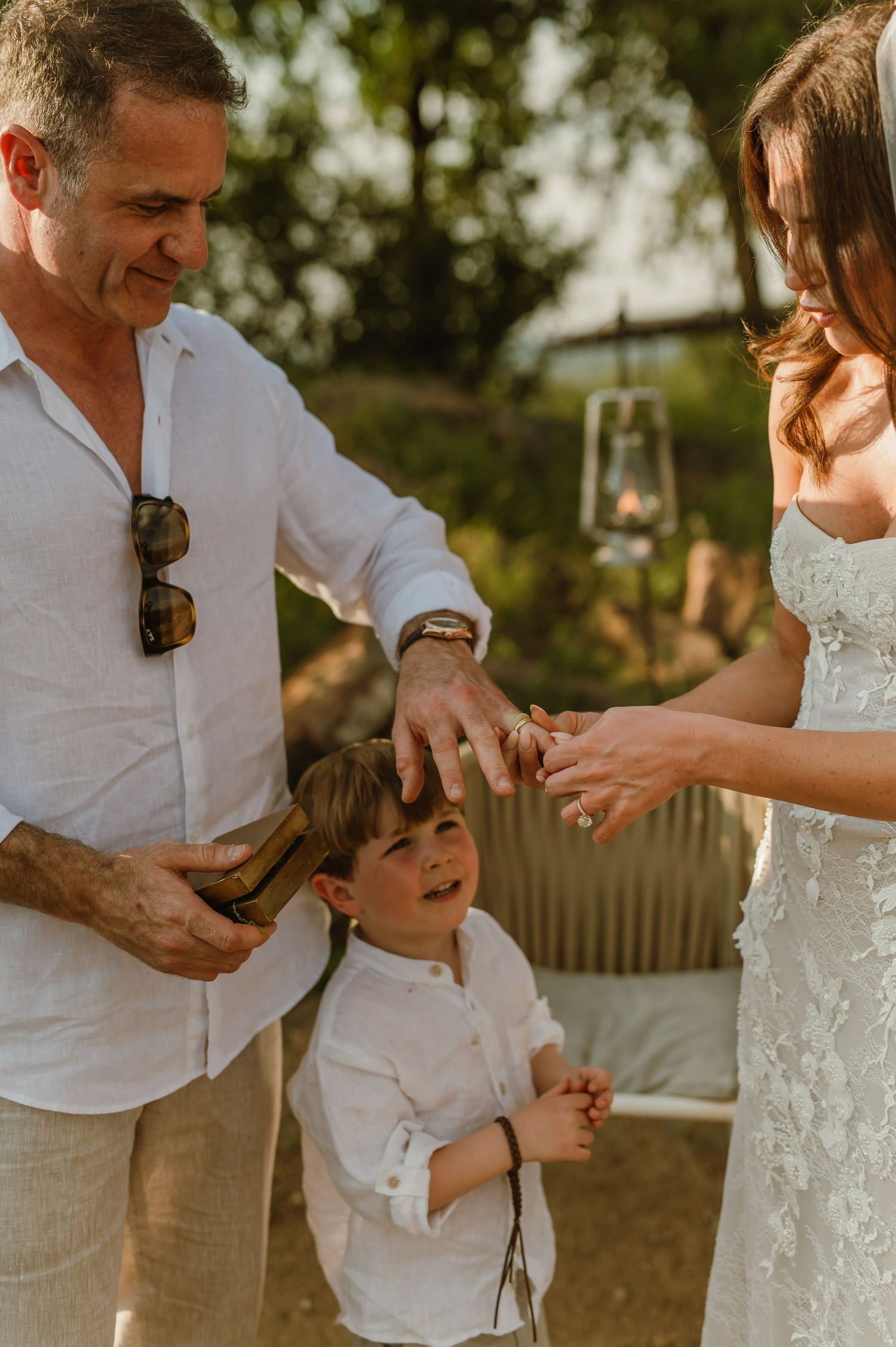 A man and woman in wedding attire share a joyful moment with a young boy outdoors at golden hour.