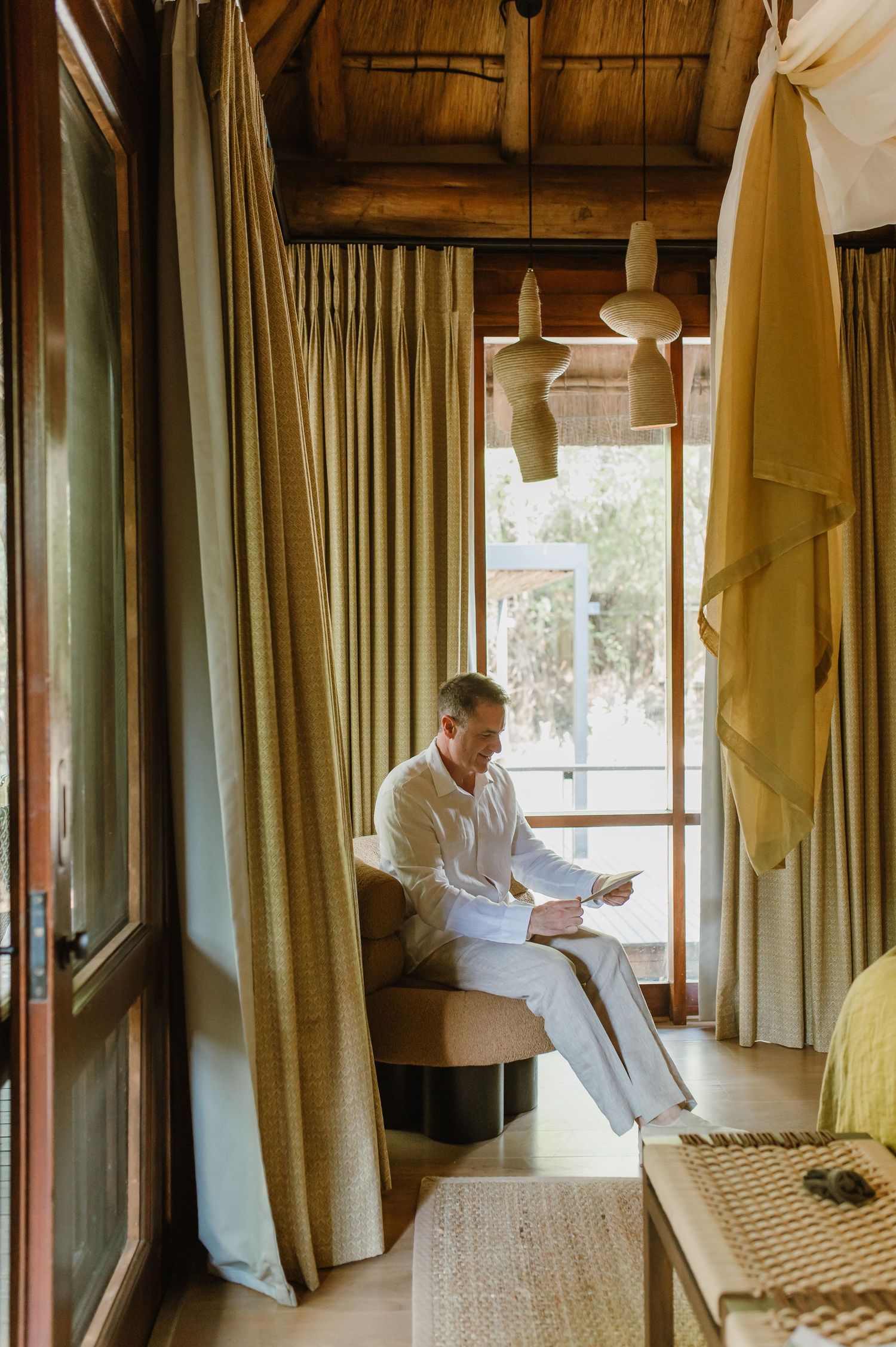 Man in white linen suit reading documents by a sunlit window in a rustic lodge room.