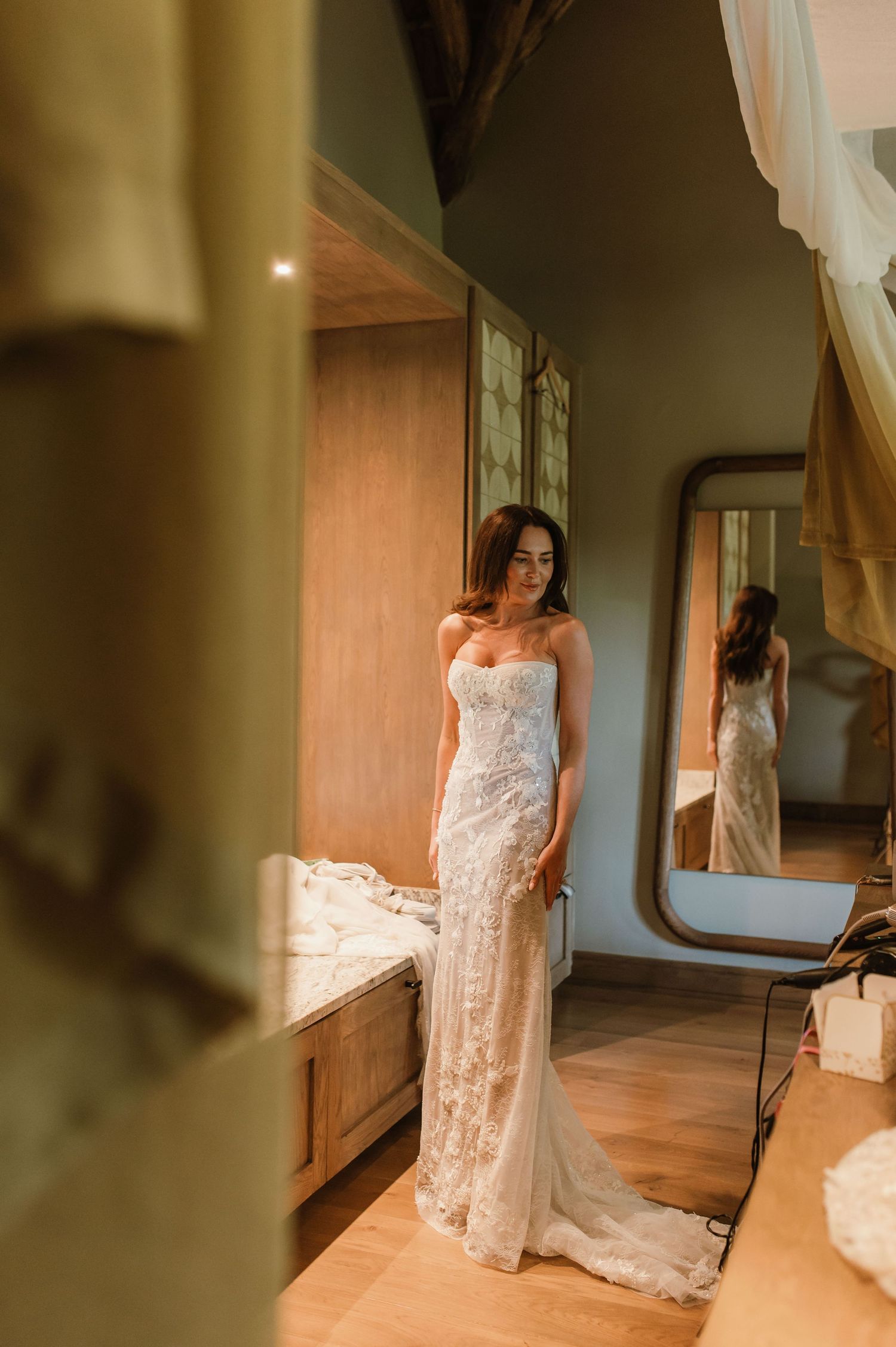 Bride in strapless lace wedding gown with train standing in rustic room reflected in oval mirror.