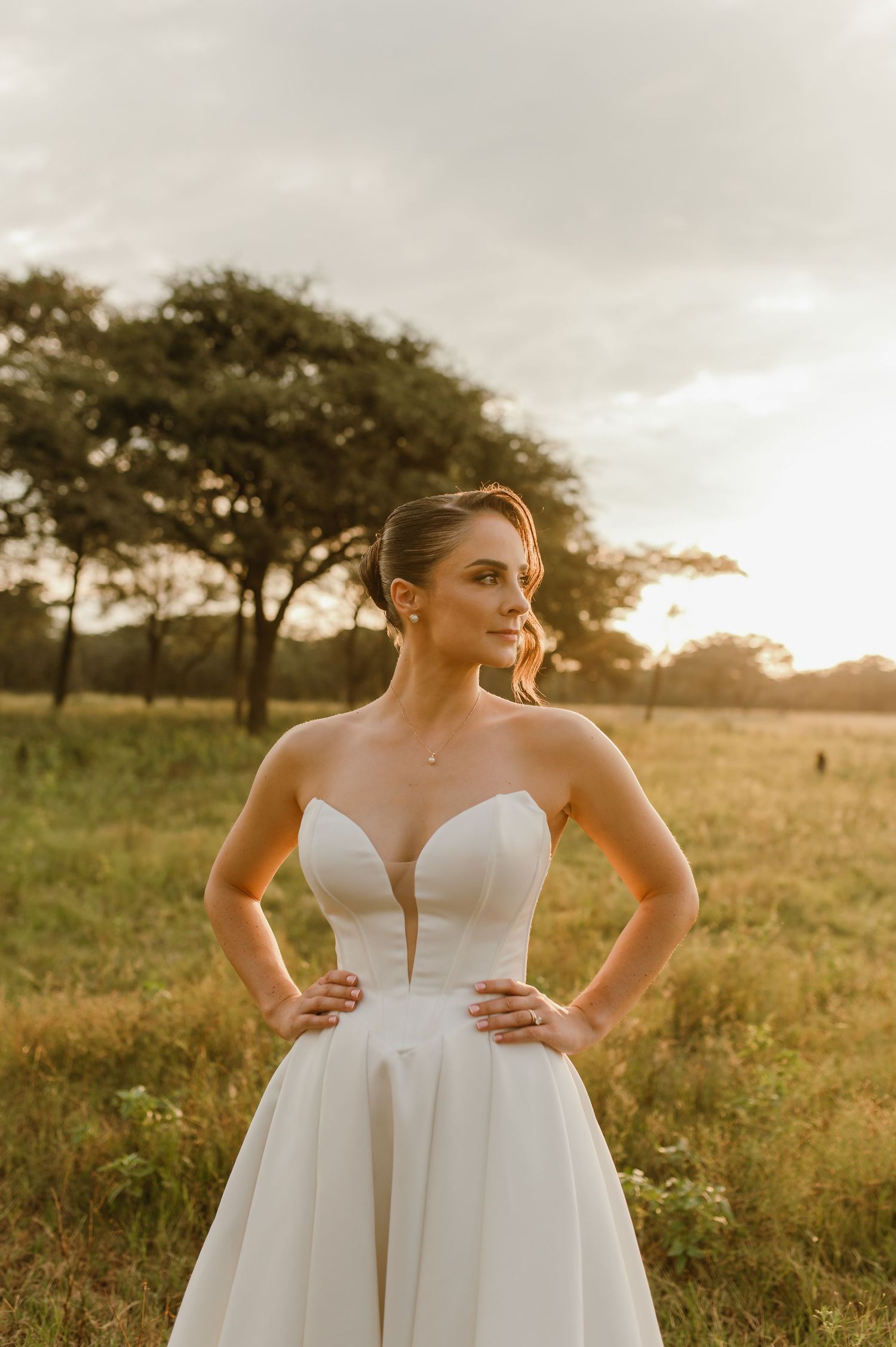 A bride in a white strapless gown poses confidently in a sun-lit field at golden hour.