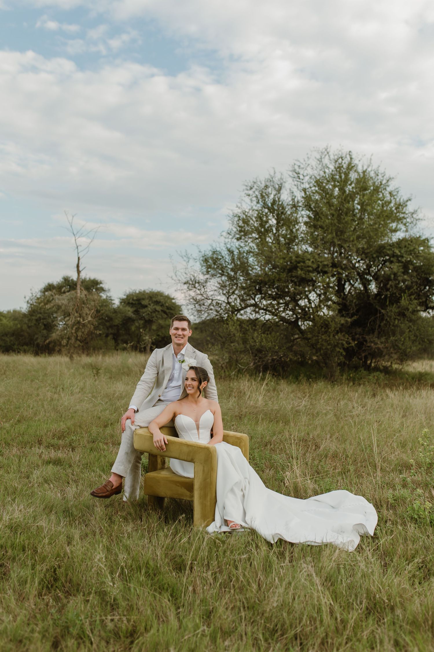 A couple in wedding attire poses together on a vintage chair in a rustic outdoor field setting.