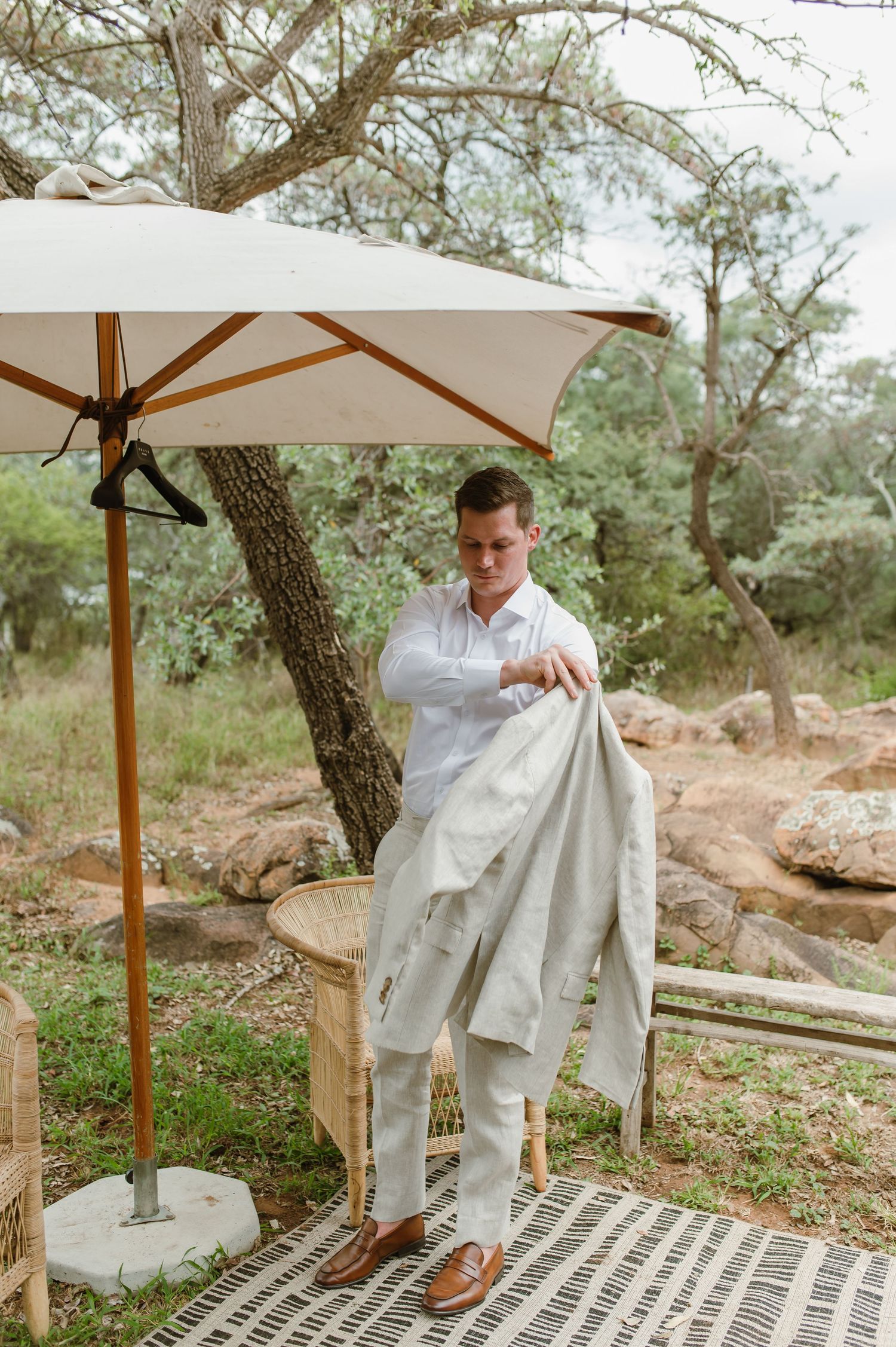Outdoor wedding setup with wooden benches and white umbrellas under natural tree canopy.