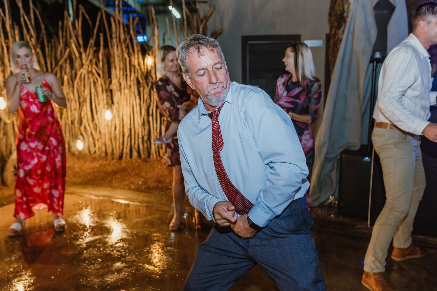 Wedding guest in blue shirt and red tie shows off energetic dance moves on reception floor.